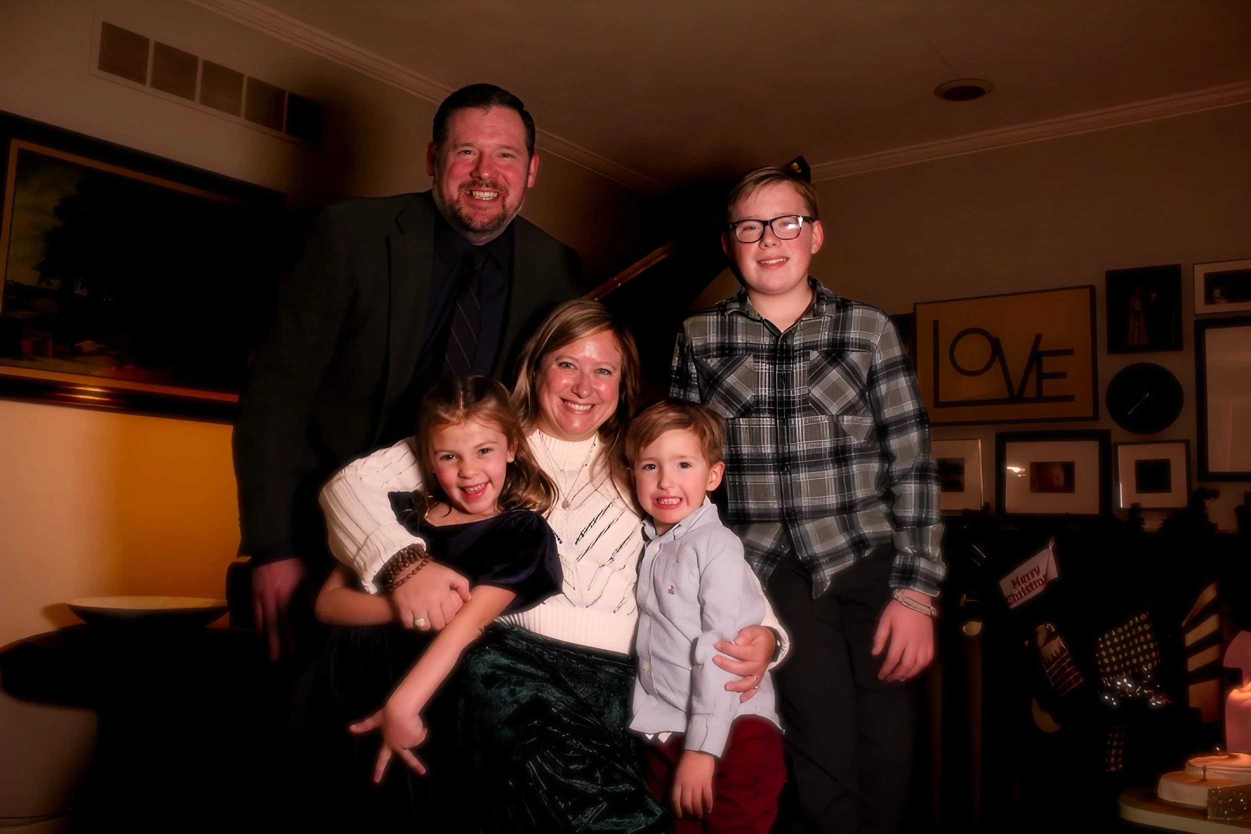 A family of five smiling and posing together indoors near a piano, with framed artwork and decor on the wall behind them.