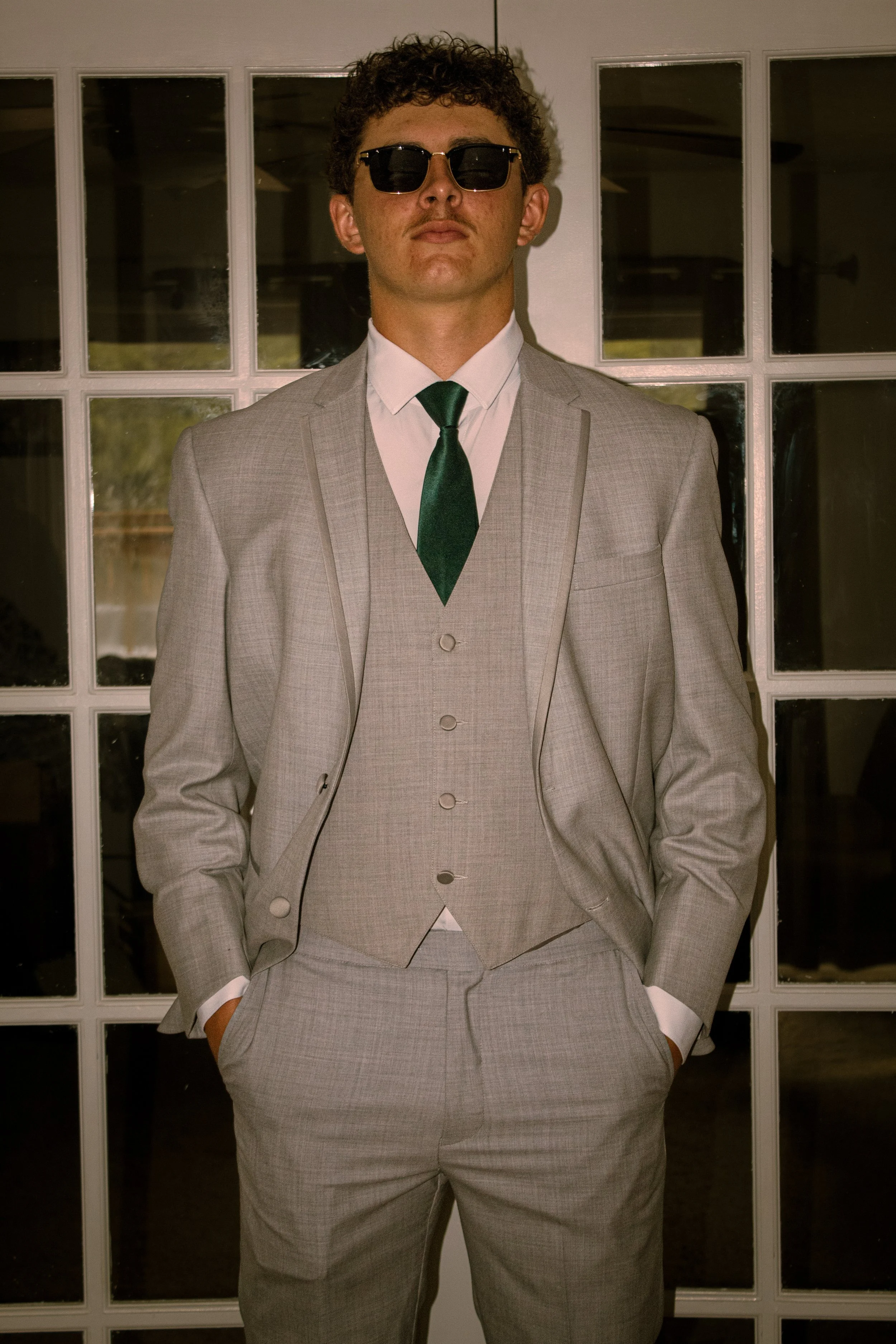 A man in a light gray suit with hands in pockets, wearing dark sunglasses, a white shirt, and a dark green tie, standing in front of a multi-paneled glass door.