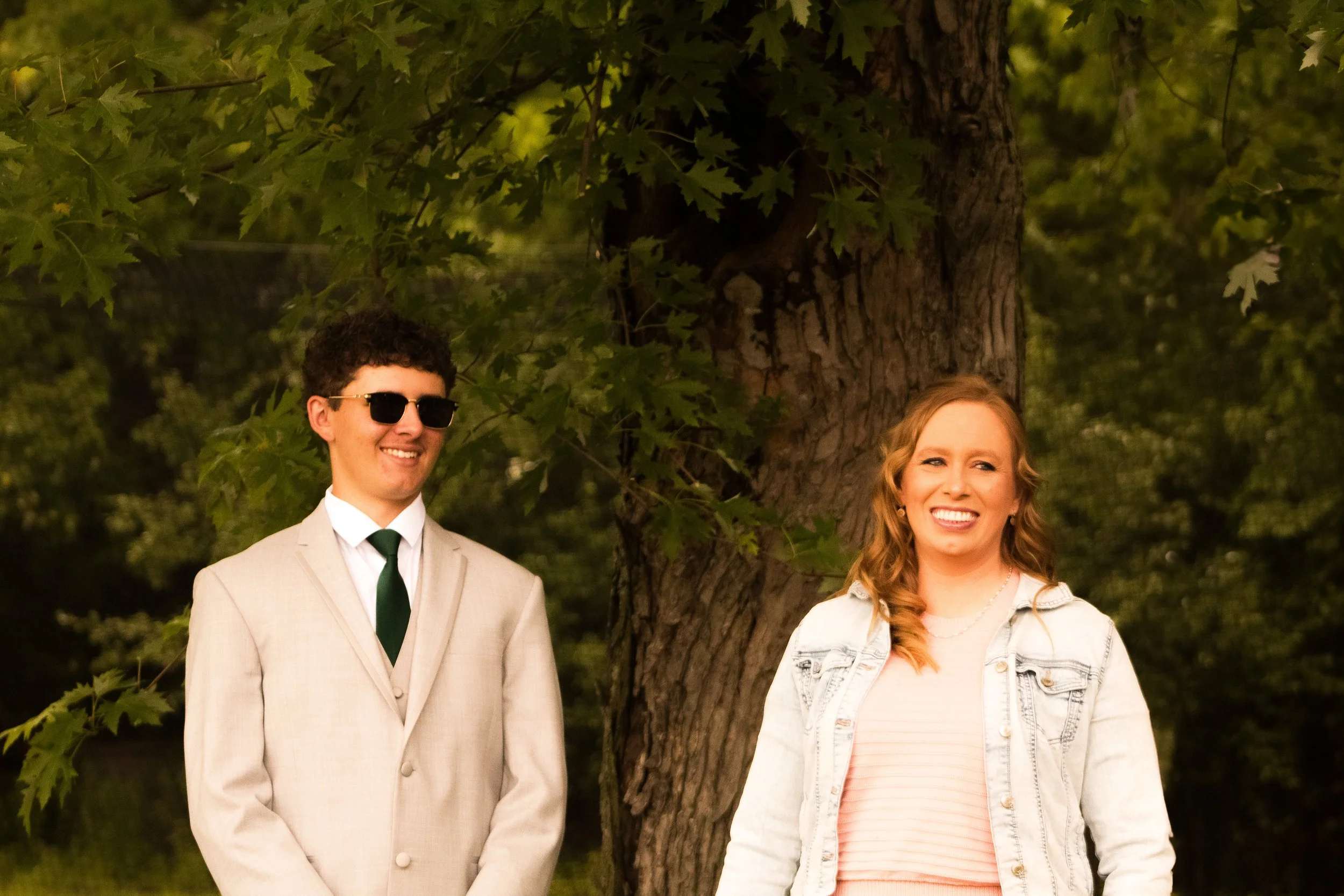 A young man in a beige suit with a white shirt, green tie, and sunglasses, standing beside a young woman with long curly hair, wearing a denim jacket over a peach striped top, both smiling outdoors next to a large tree with green leaves.