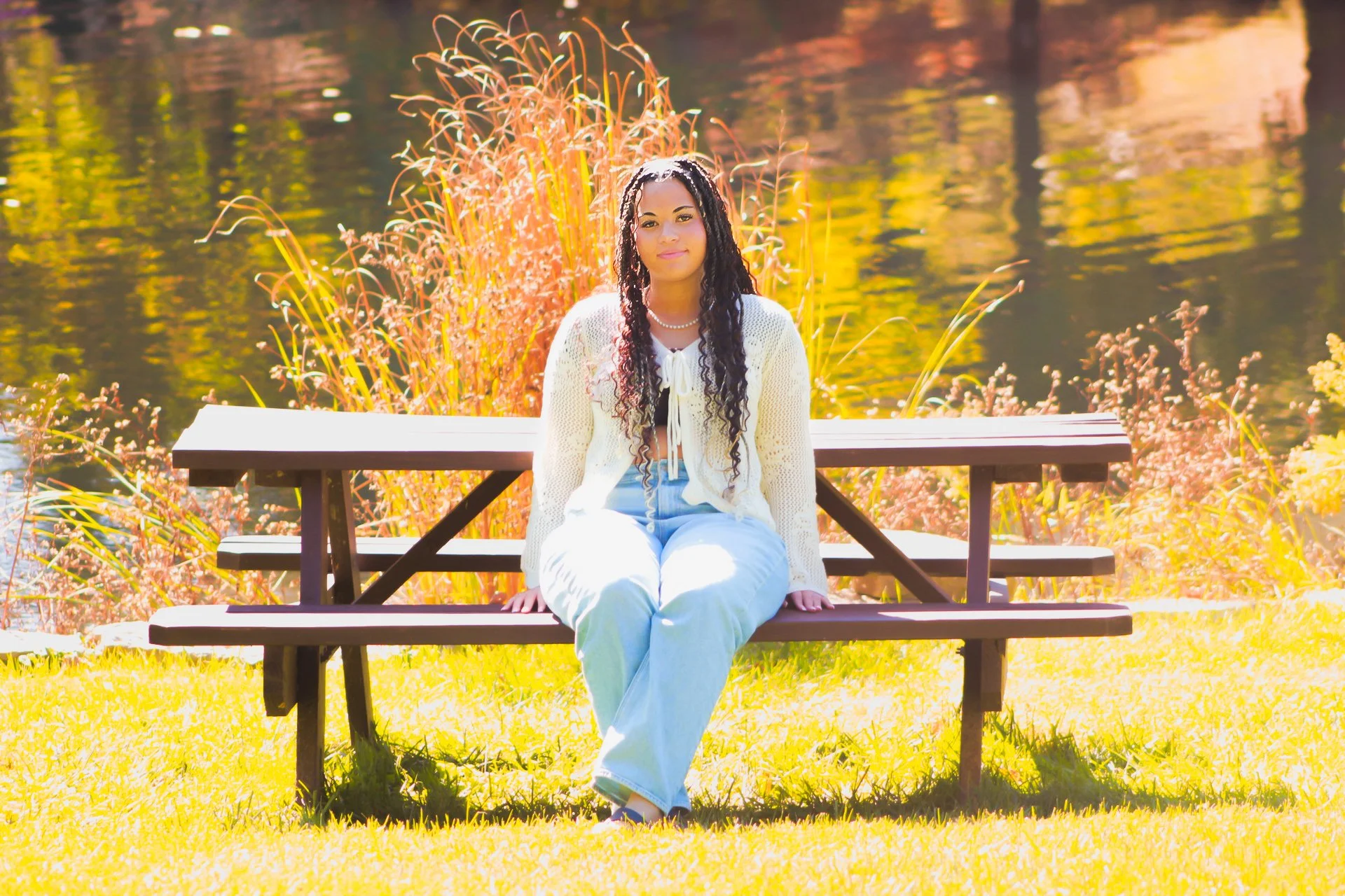 Woman in a white cardigan and light blue pants sitting on a picnic table near a body of water on a sunny day, surrounded by autumn foliage.