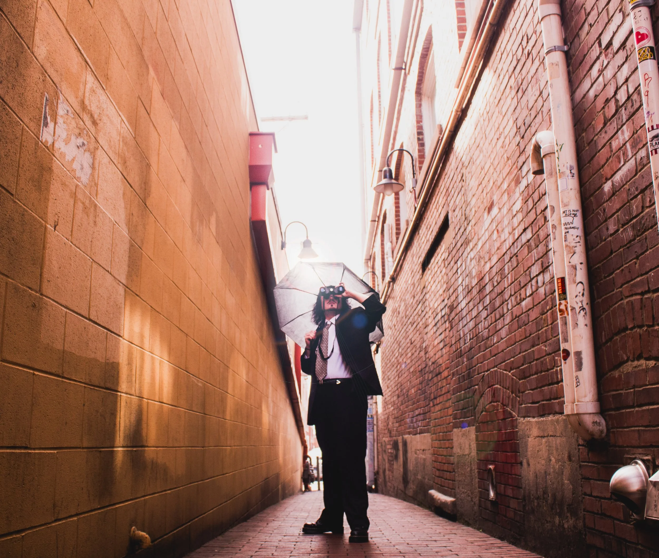 Man in black suit holding an umbrella and taking a photo in an alleyway with brick walls.