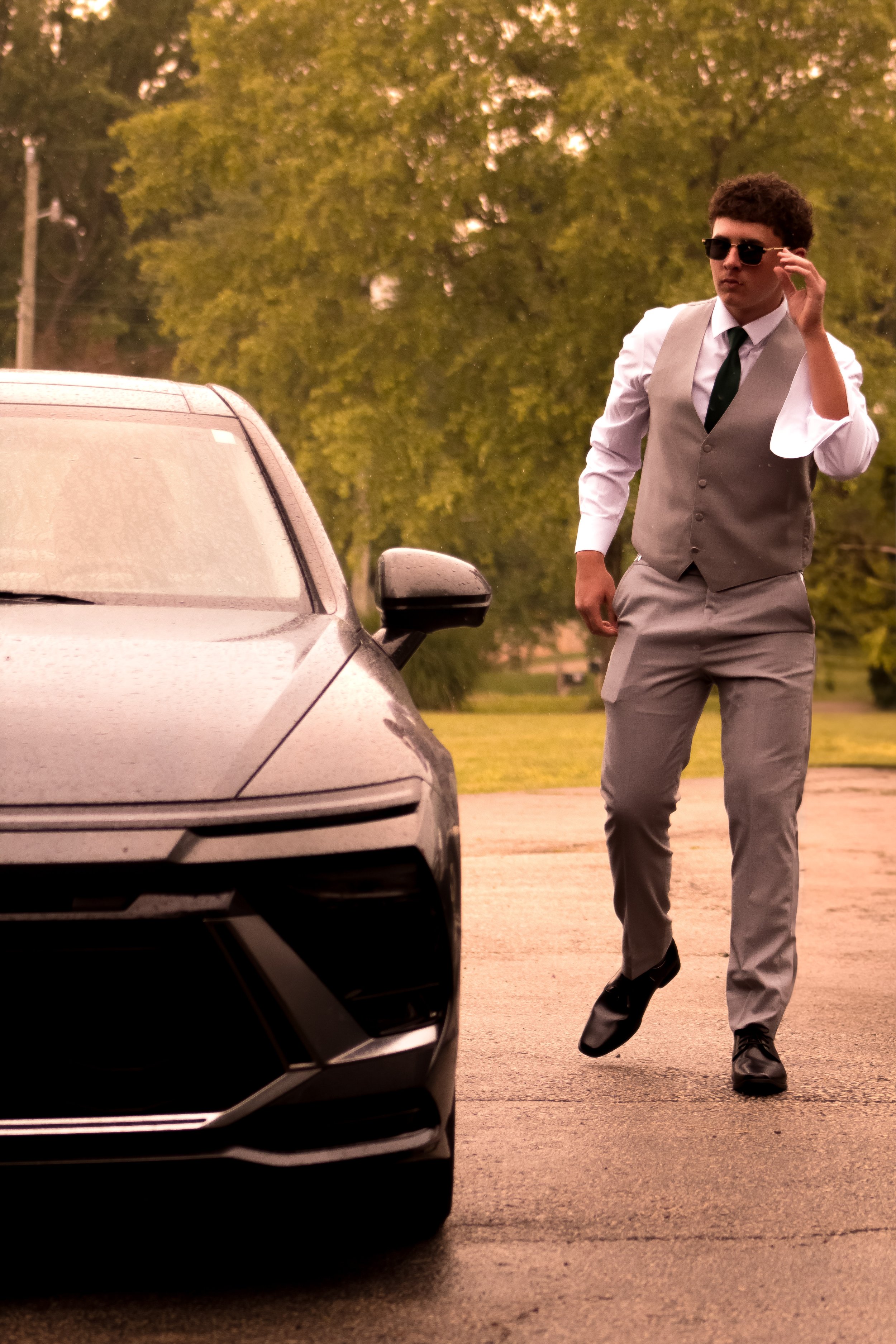 A young man in a white shirt, gray vest, gray trousers, black shoes, and sunglasses walks in a parking lot next to a silver car with a black front grill, and green trees in the background.