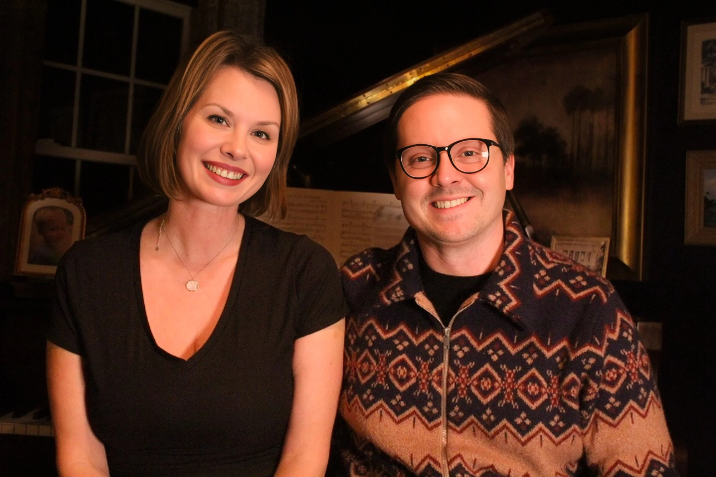 A woman with short brown hair and a man with glasses sitting together in a warmly lit room, smiling at the camera. The woman is wearing a black dress and the man is wearing a patterned sweater.
