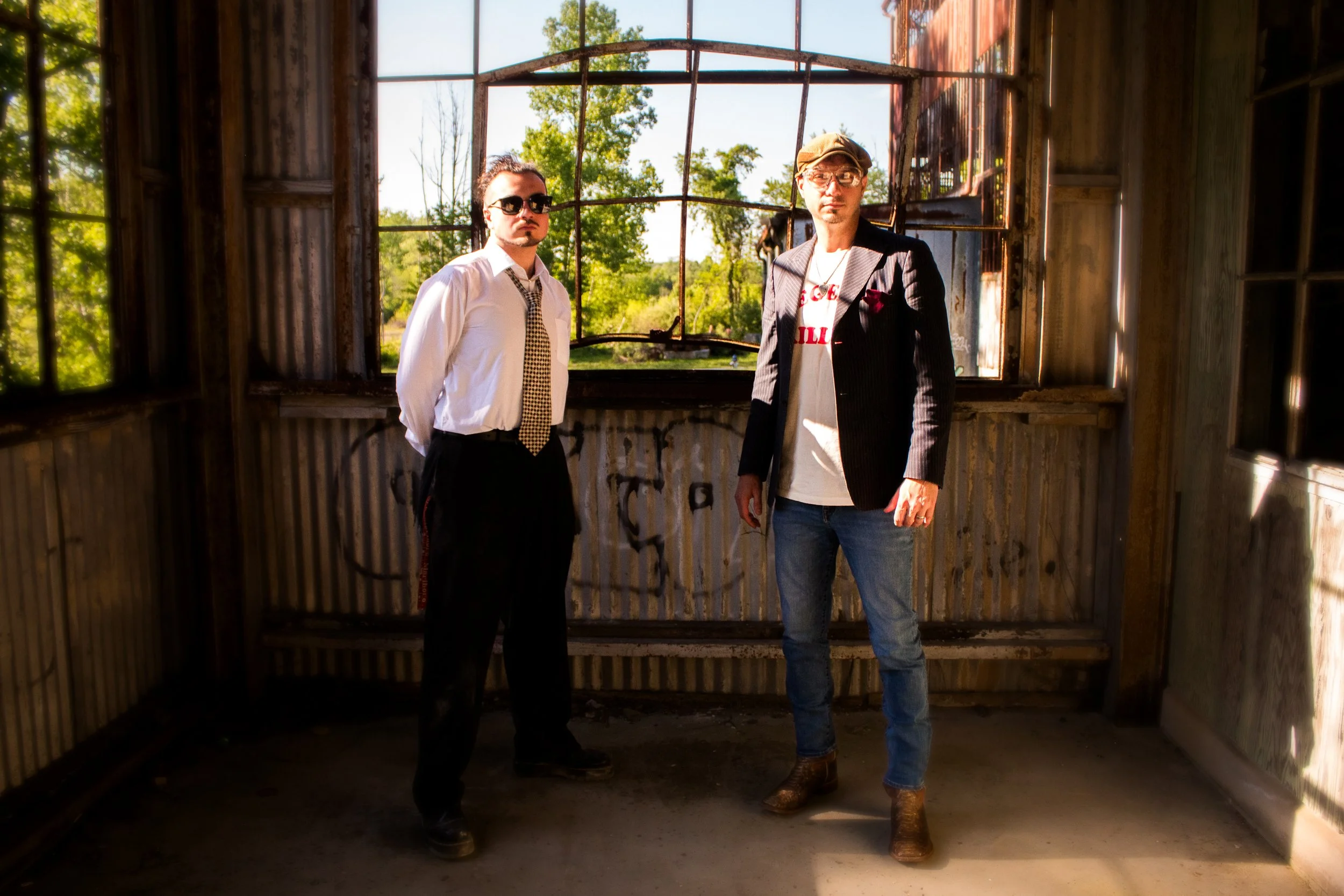 Two men standing inside an abandoned, rusty, wooden structure with large open windows, trees and blue sky visible outside.