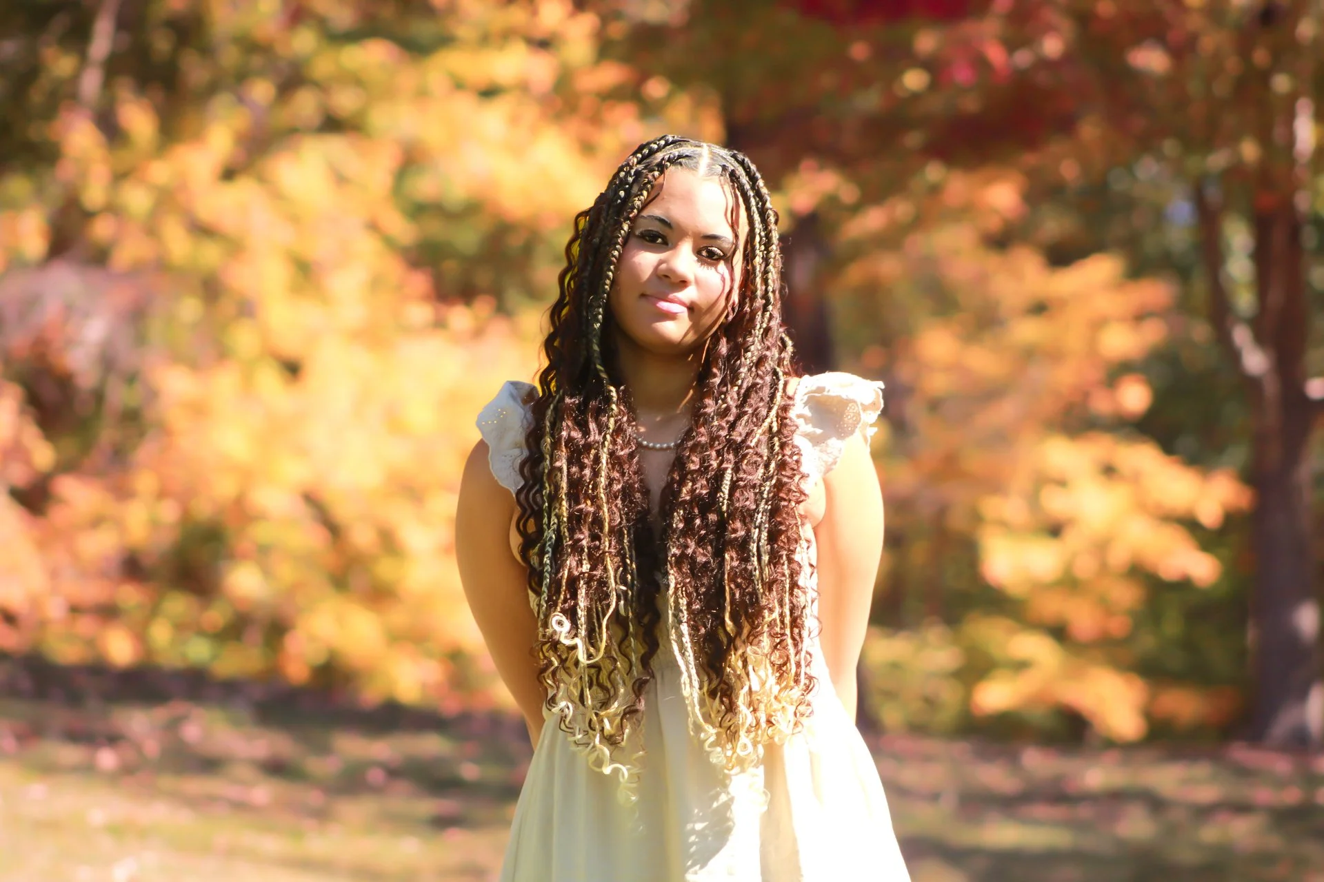 A woman with long, curly, multicolored hair standing outdoors in a park during autumn, with trees in the background showing fall foliage.