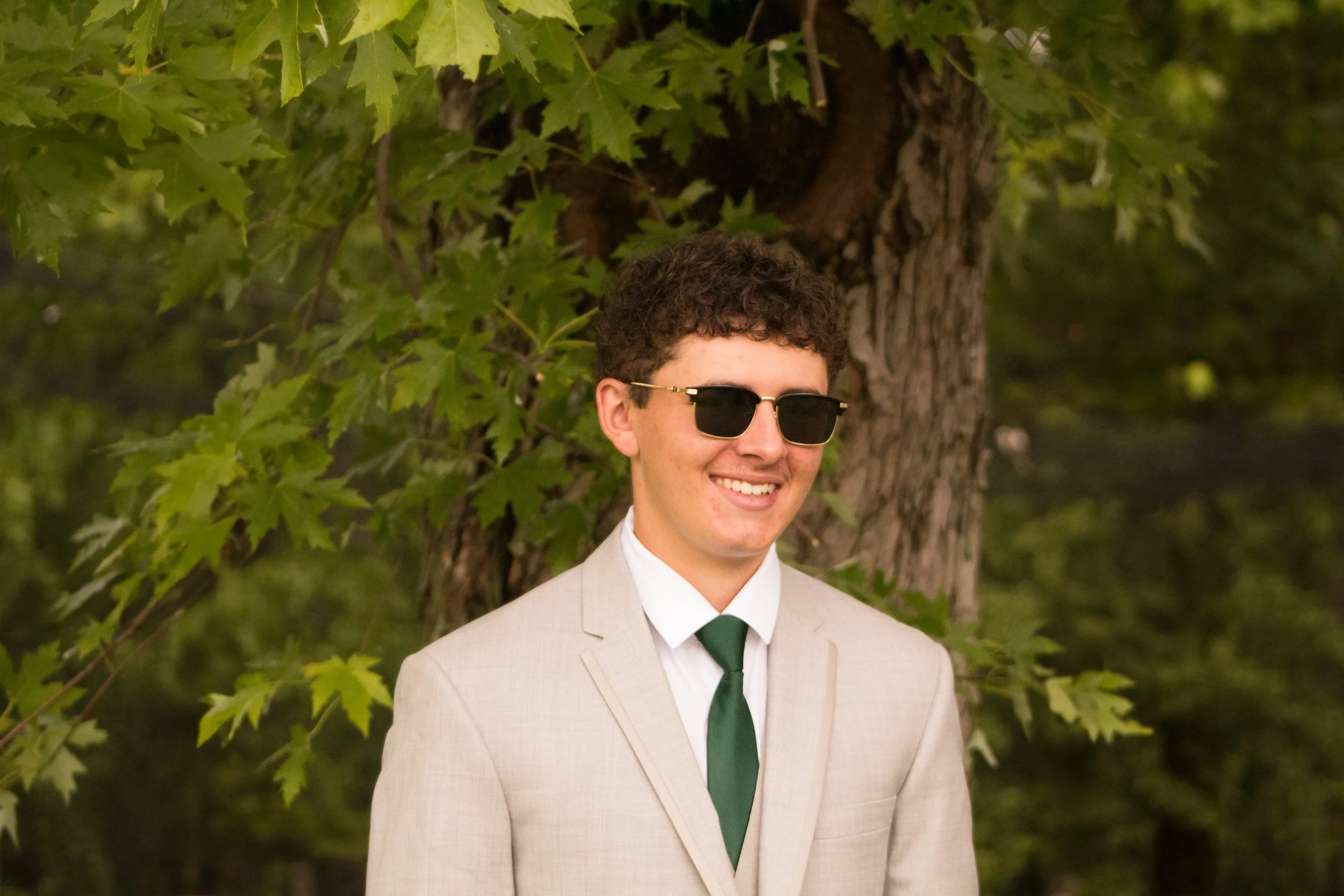 A young man with curly hair wearing a beige suit, white shirt, dark green tie, sunglasses, smiling outdoors in front of a large tree with green leaves.