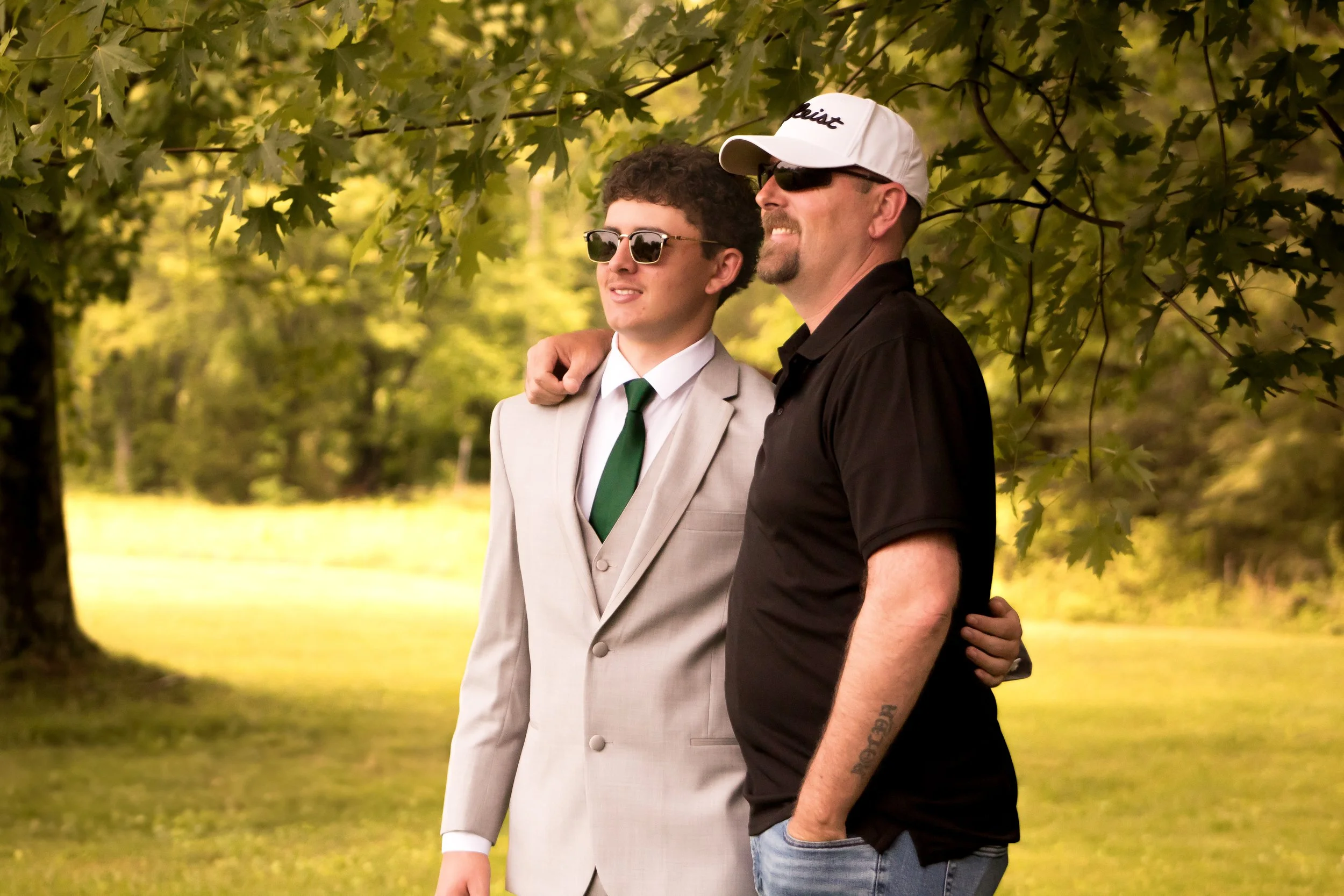 Two men are standing outdoors under a tree, smiling and with their arms around each other's shoulders. The man on the left is wearing a beige suit, white shirt, green tie, and sunglasses. The man on the right is wearing a black shirt, a white cap, su