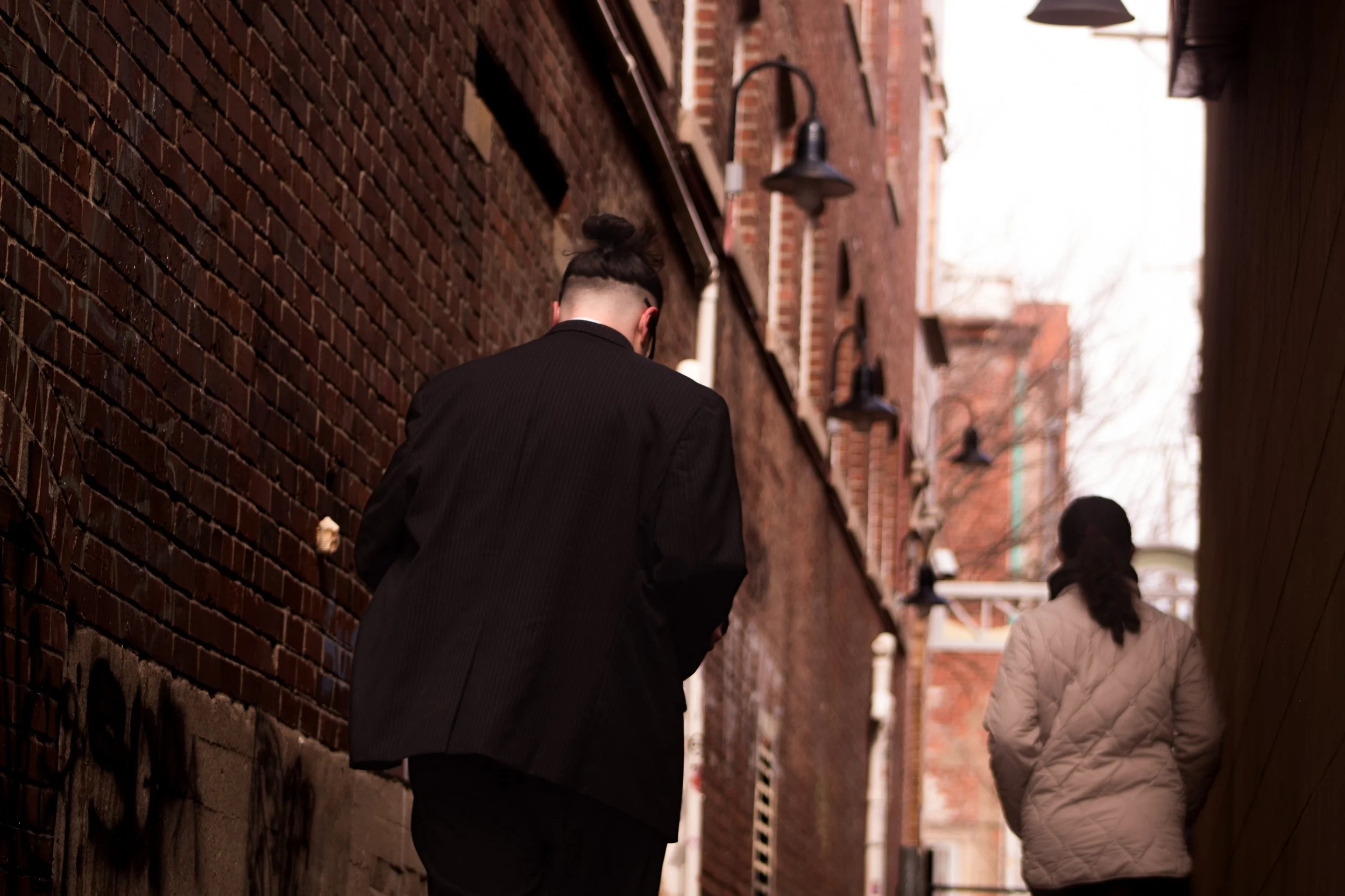 Two people walking down a narrow alleyway with brick walls and black wall-mounted lights, one wearing a black jacket with a high bun hairstyle, the other in a beige quilted jacket with long dark hair.