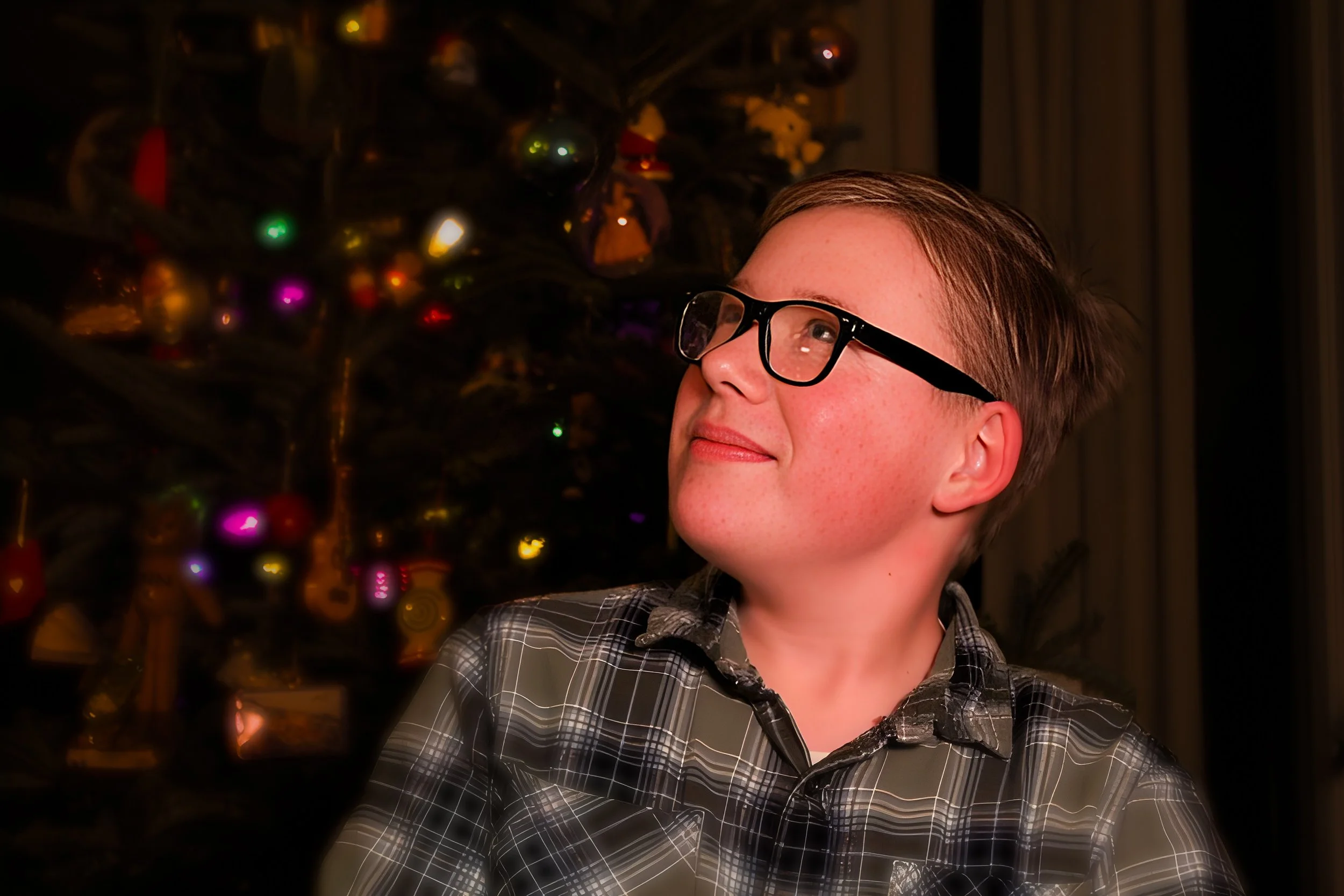 A boy with glasses and a plaid shirt looking to the side in front of a decorated Christmas tree.