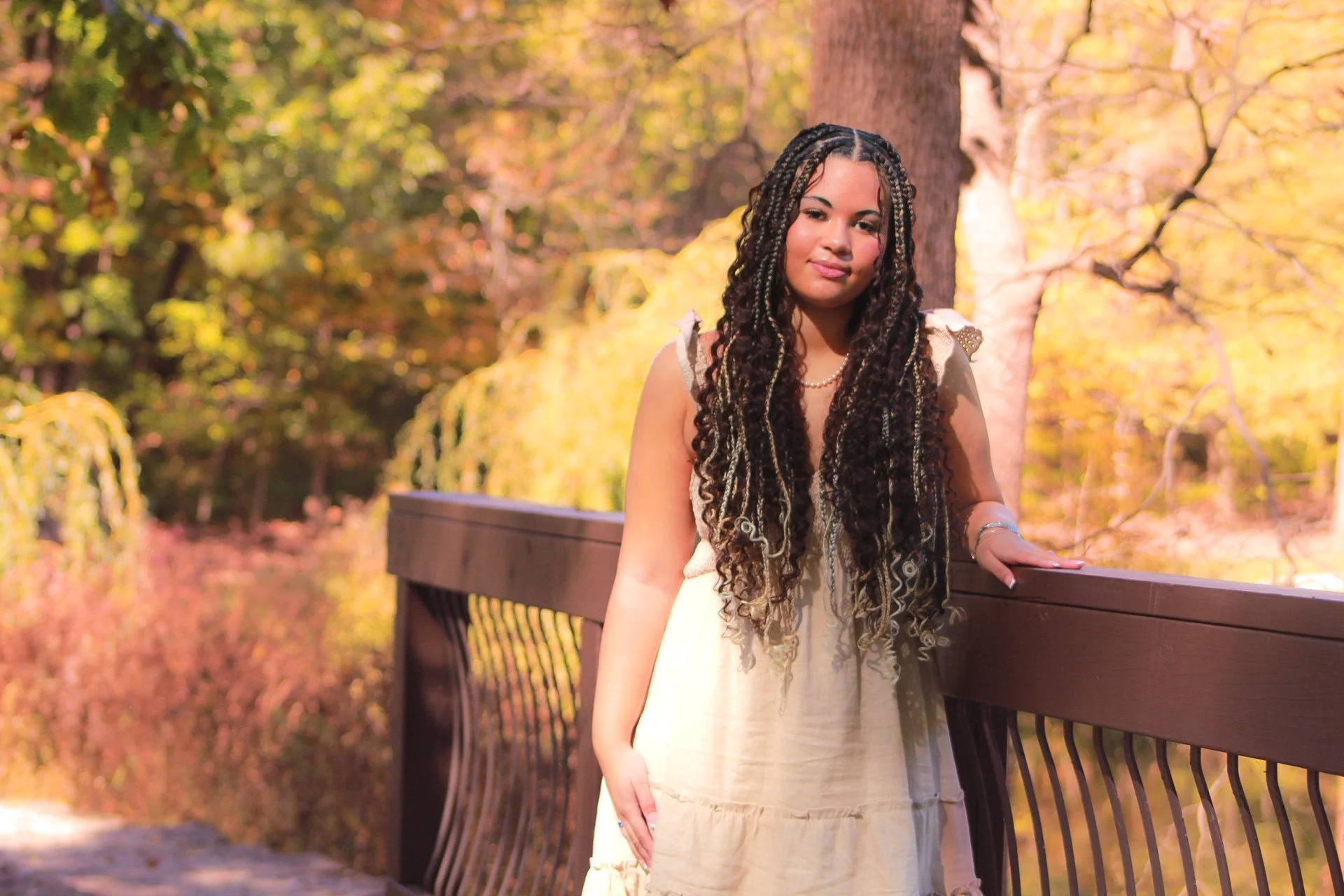 A woman with long braids, wearing a cream dress, stands on a wooden bridge with fall-colored trees in the background.