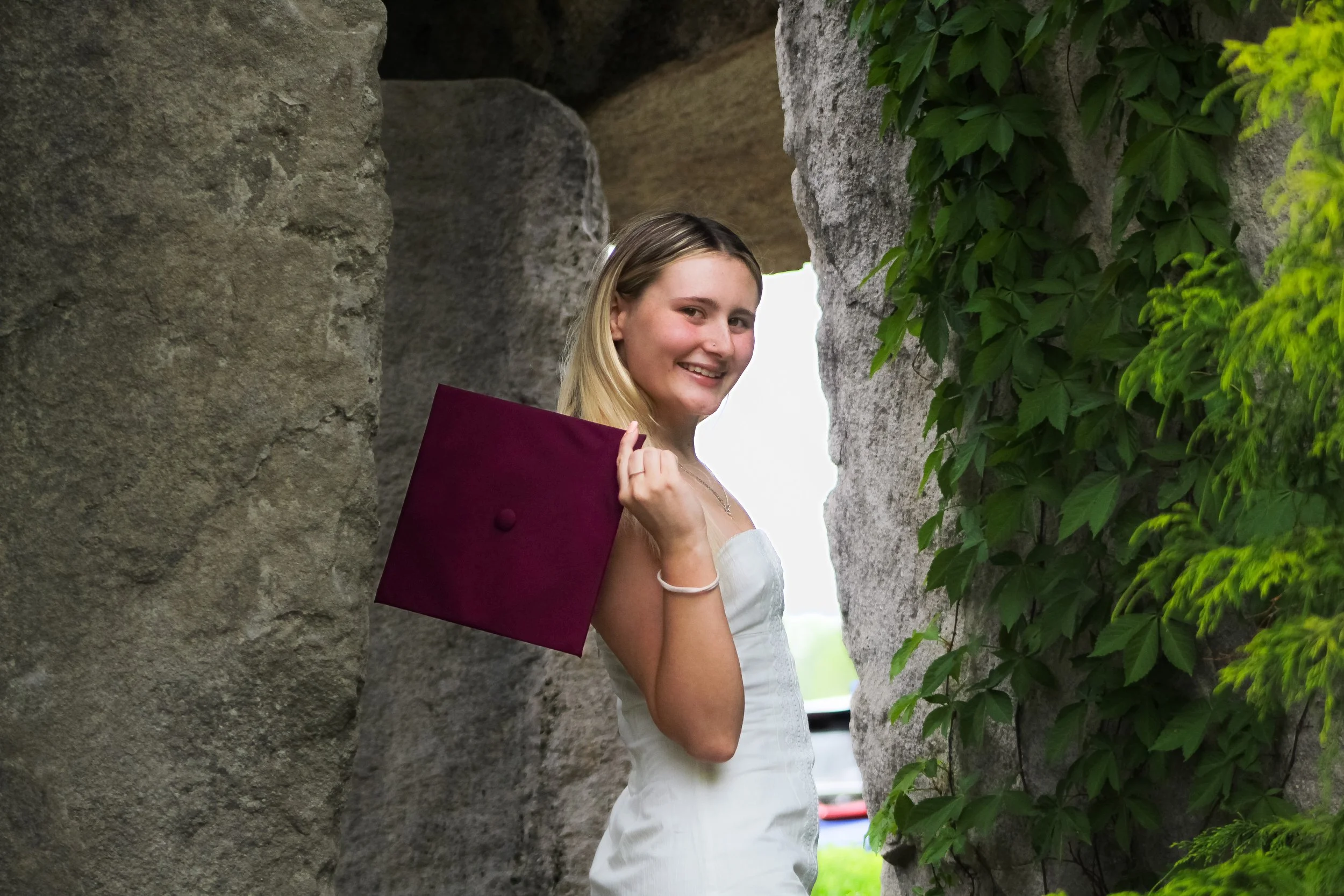 A young woman in a white dress smiling at the camera, standing between two large stone rocks with green ivy on the right side.
