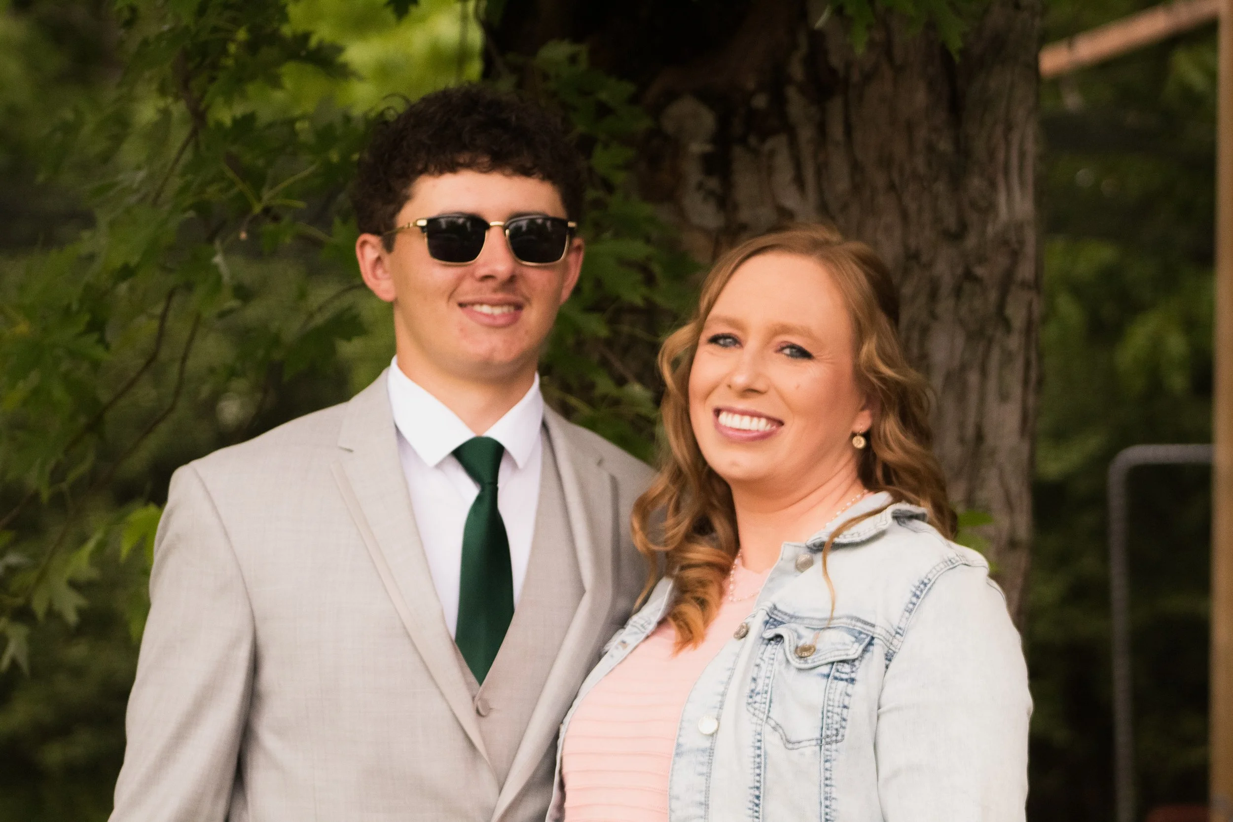 Two women smiling, standing outdoors near a tree, with greenery in the background. One woman has curly hair and is wearing a denim jacket and a pink shirt; the other has straight hair and is wearing a light-colored blazer, sunglasses, and a white shi