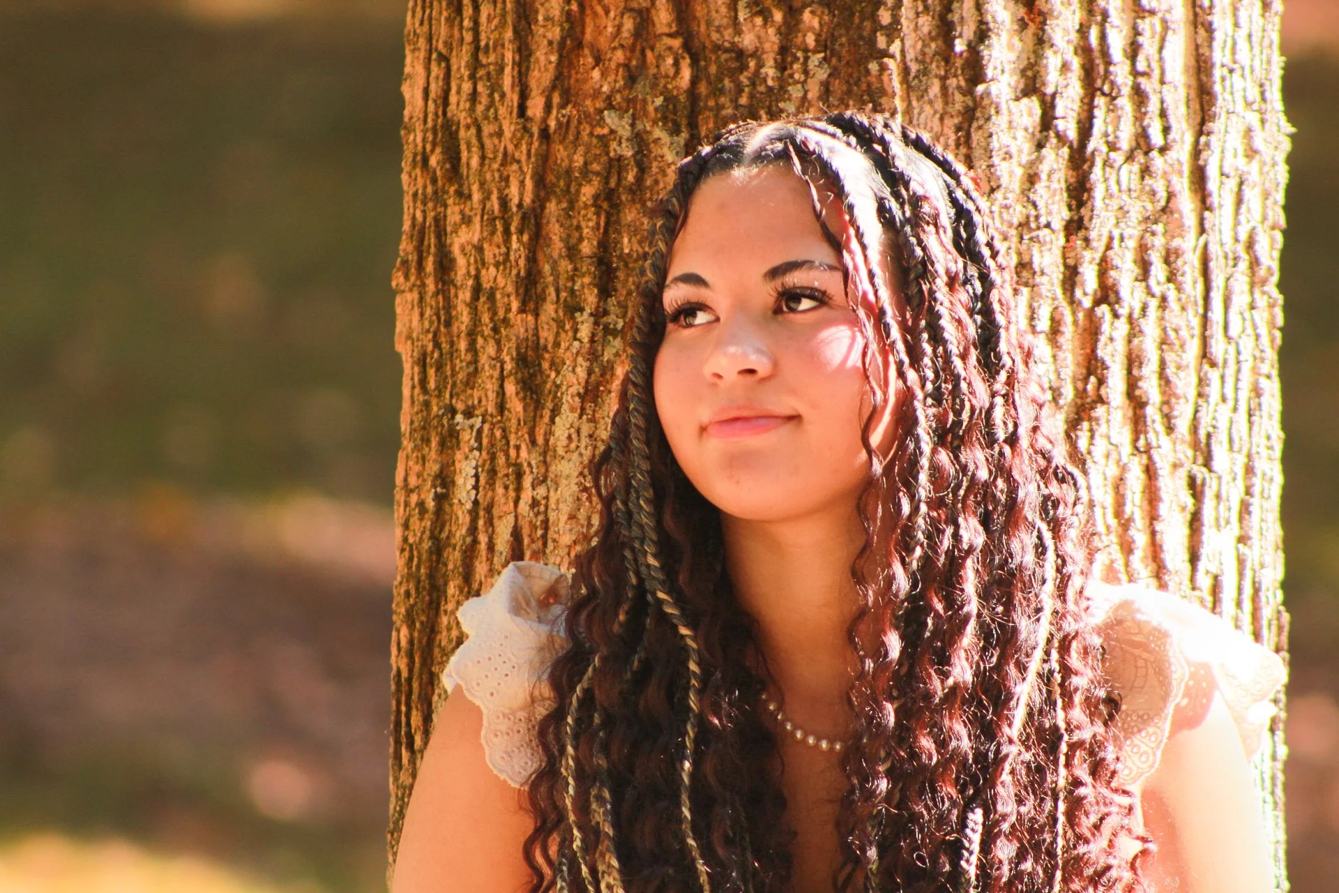 A young woman with braided hair, wearing a white lace top and a pearl necklace, sitting outdoors against a tree, looking away with a slight smile.