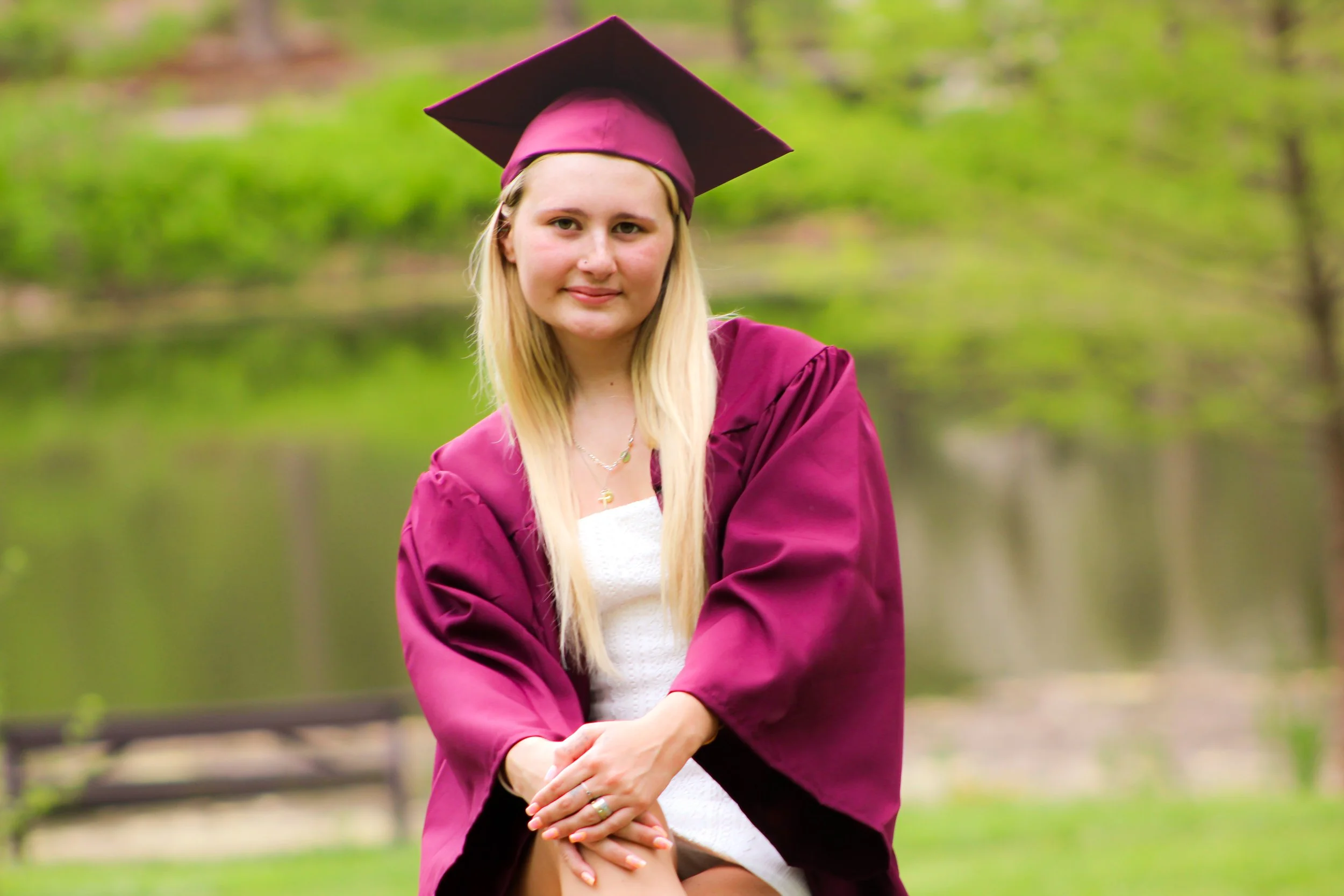 A young woman in a maroon graduation gown and cap, sitting outdoors near a body of water with green trees in the background, smiling softly.