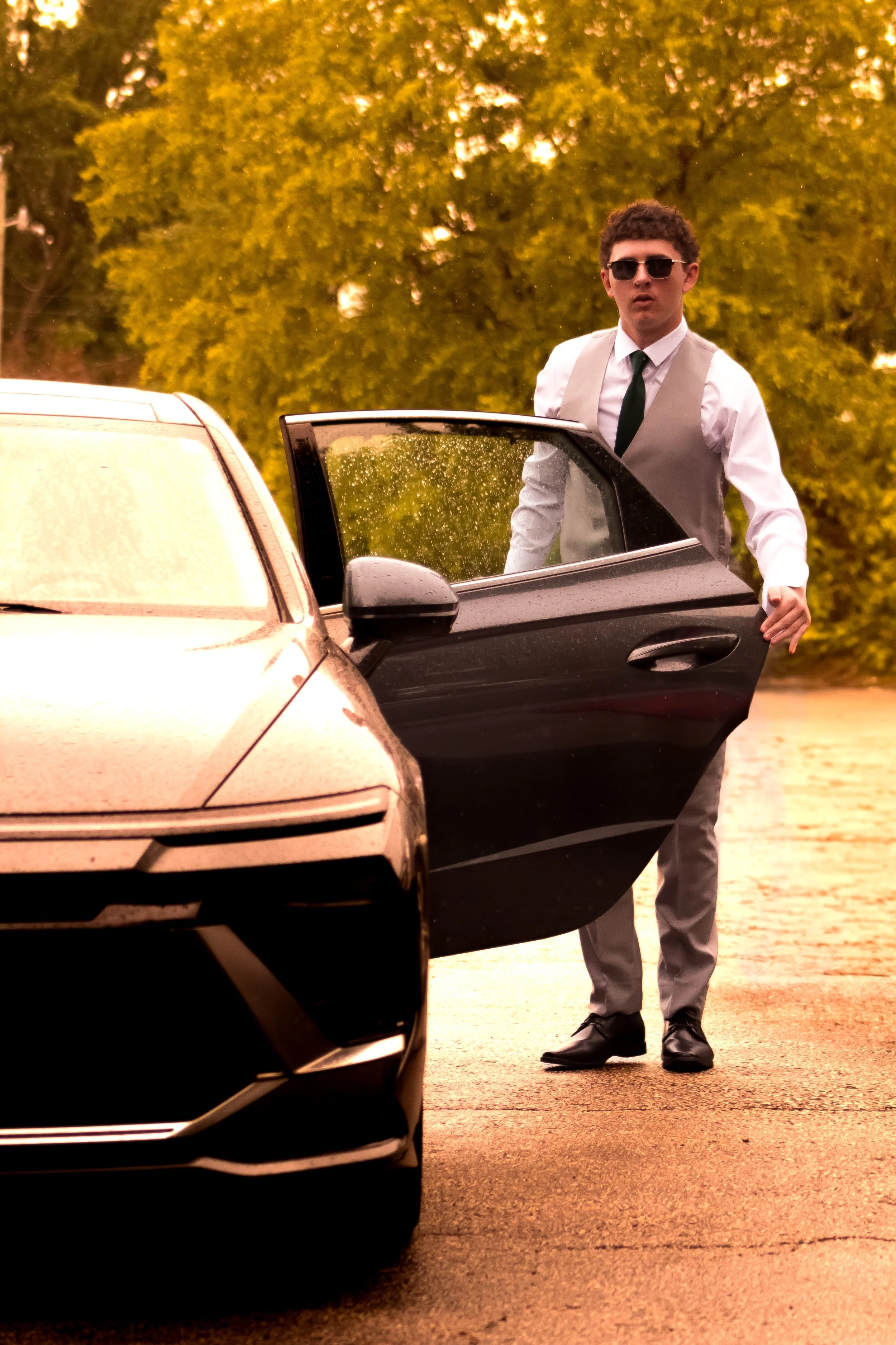 A young man in a light gray suit with a white shirt, black tie, and sunglasses stands outside near a black car, holding the open passenger door. The background features trees with yellow-green leaves, suggesting autumn.