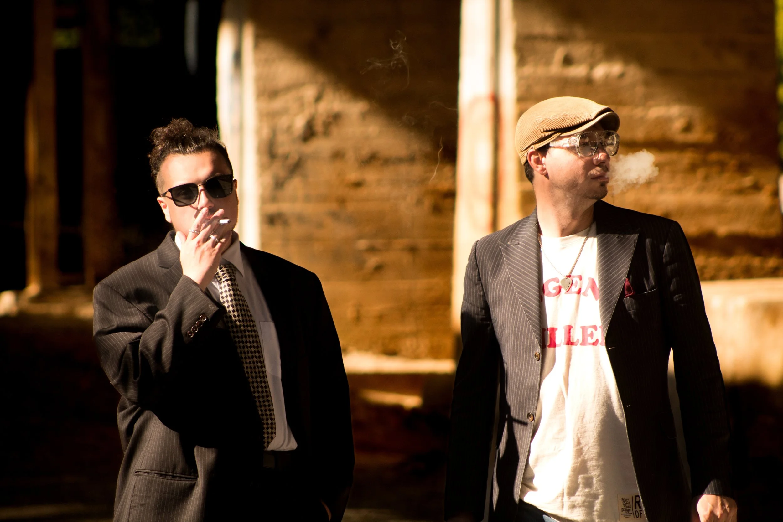 Two men in suits and sunglasses standing outdoors near a wooden structure. One is smoking, and the other has steam or vapor coming from his mouth. Both are dressed stylishly, and the scene has warm, natural lighting.