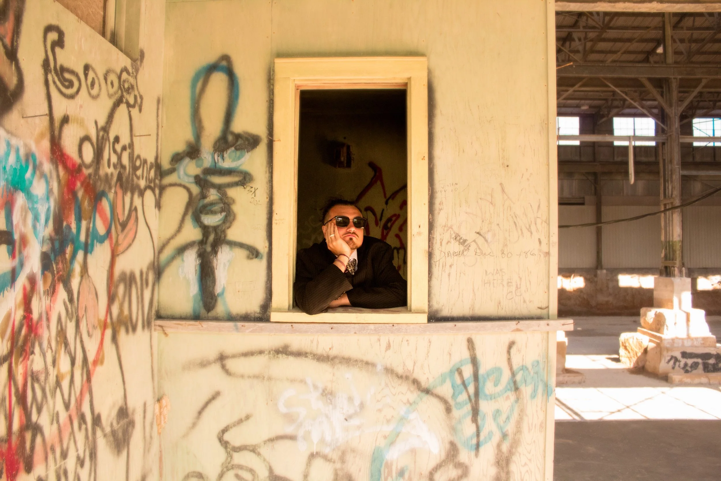 A man wearing sunglasses and a dark jacket is leaning on a window sill with his hand resting on his face inside an unfinished, graffiti-covered building.