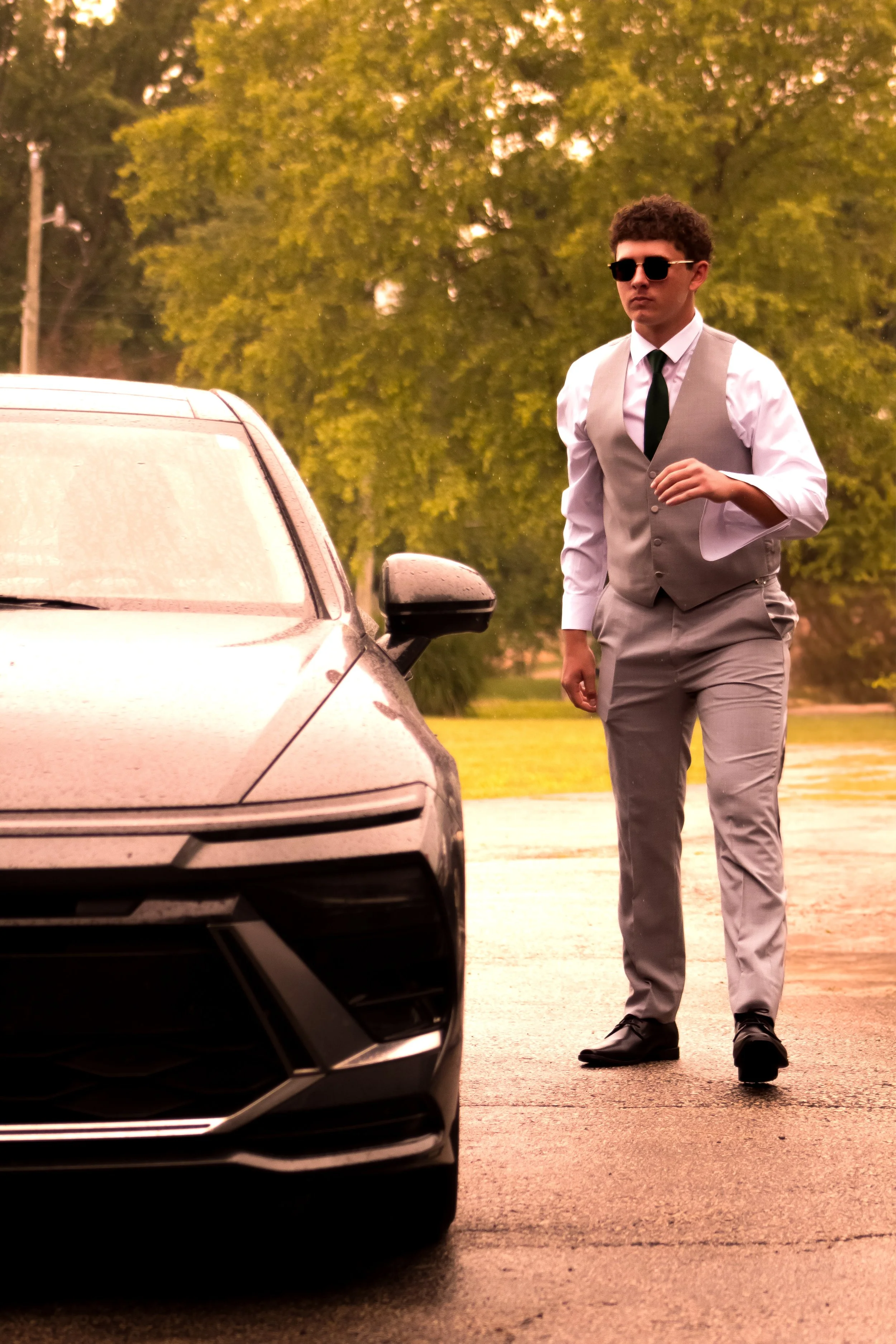 A young man in a tan vest, white shirt, black tie, and sunglasses walking near a black car in an outdoor setting with green trees in the background.