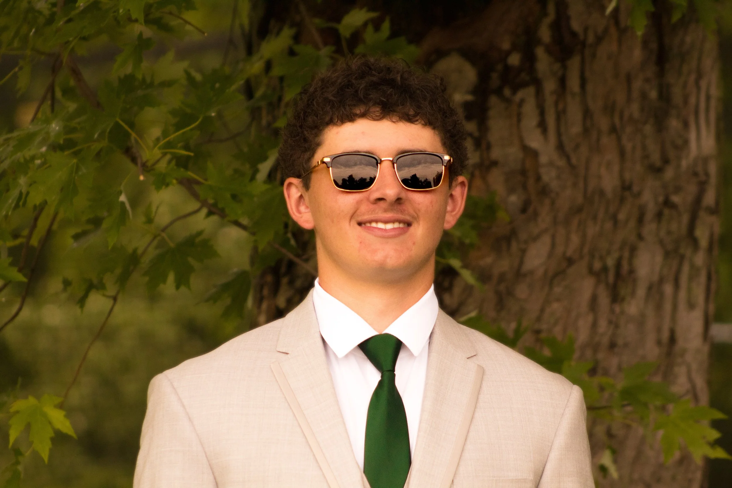 Young man in a beige suit, white shirt, and black tie, wearing sunglasses, smiling, standing outdoors near a large tree with green leaves.