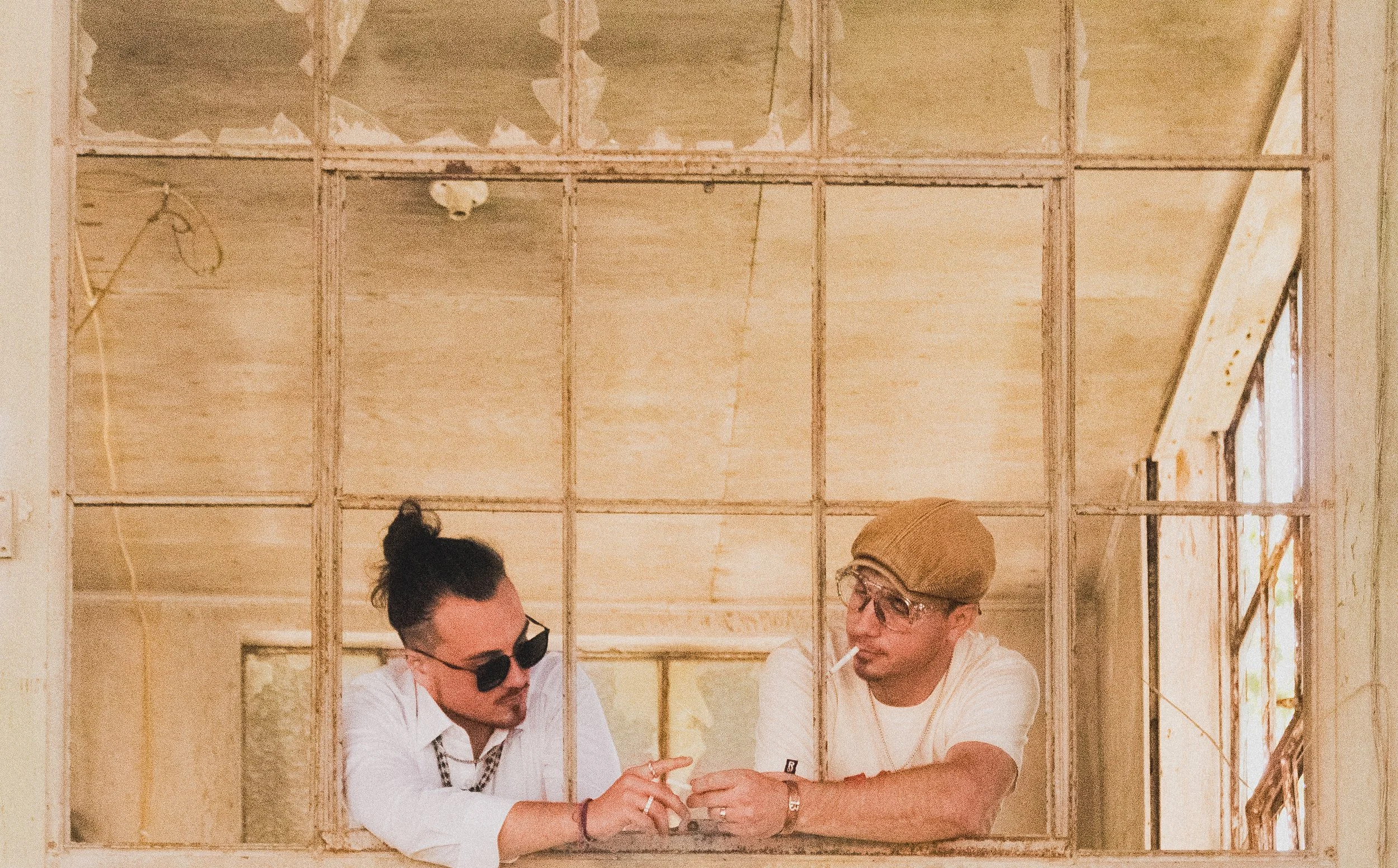 Two men working on a construction or renovation project inside a wooden frame wall, with one person wearing glasses and a beanie, and the other with dark hair tied up, both engaged in conversation.