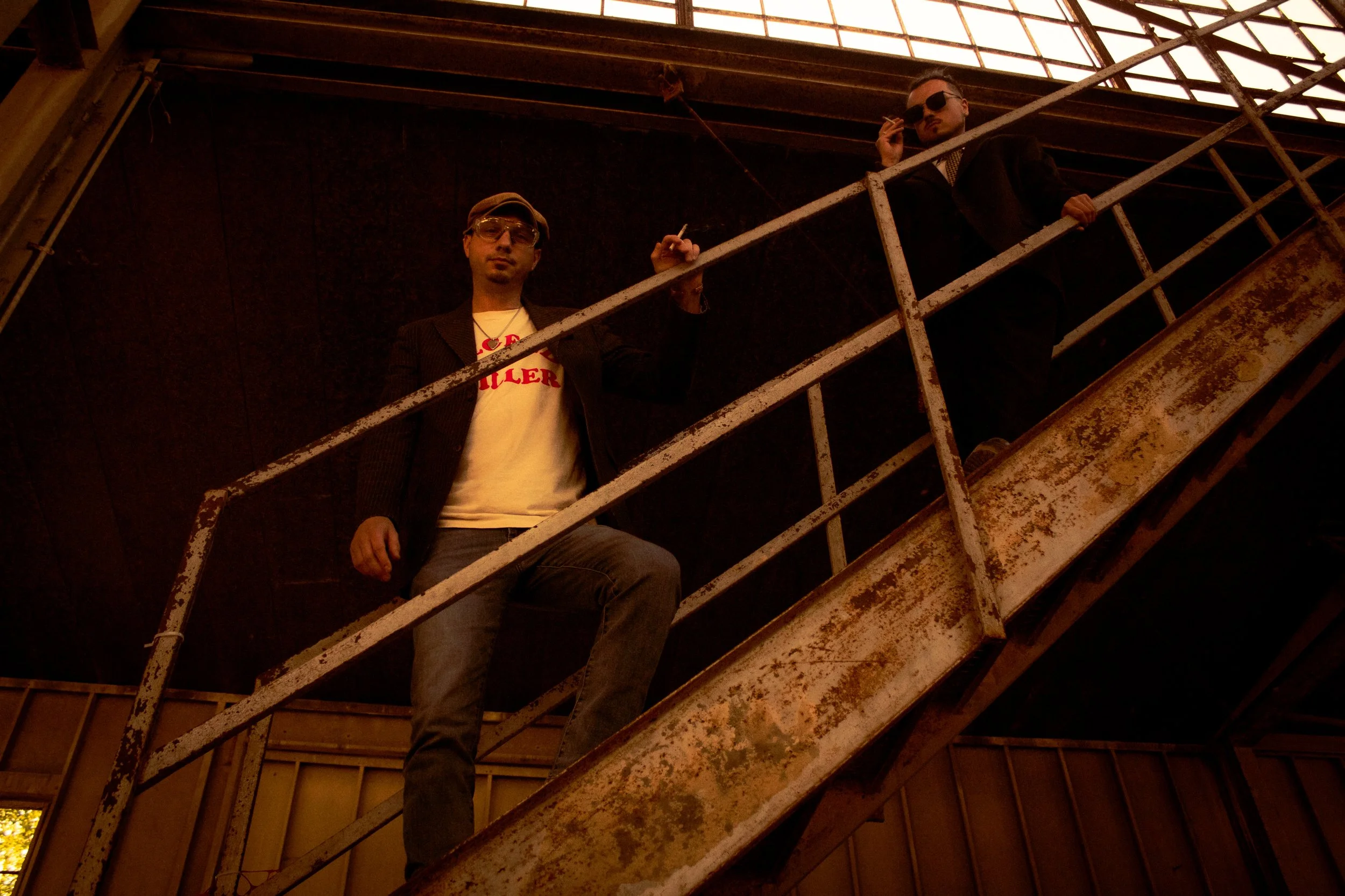 Two men standing on a rusty metal staircase inside an unfinished building with wooden framing and large windows.