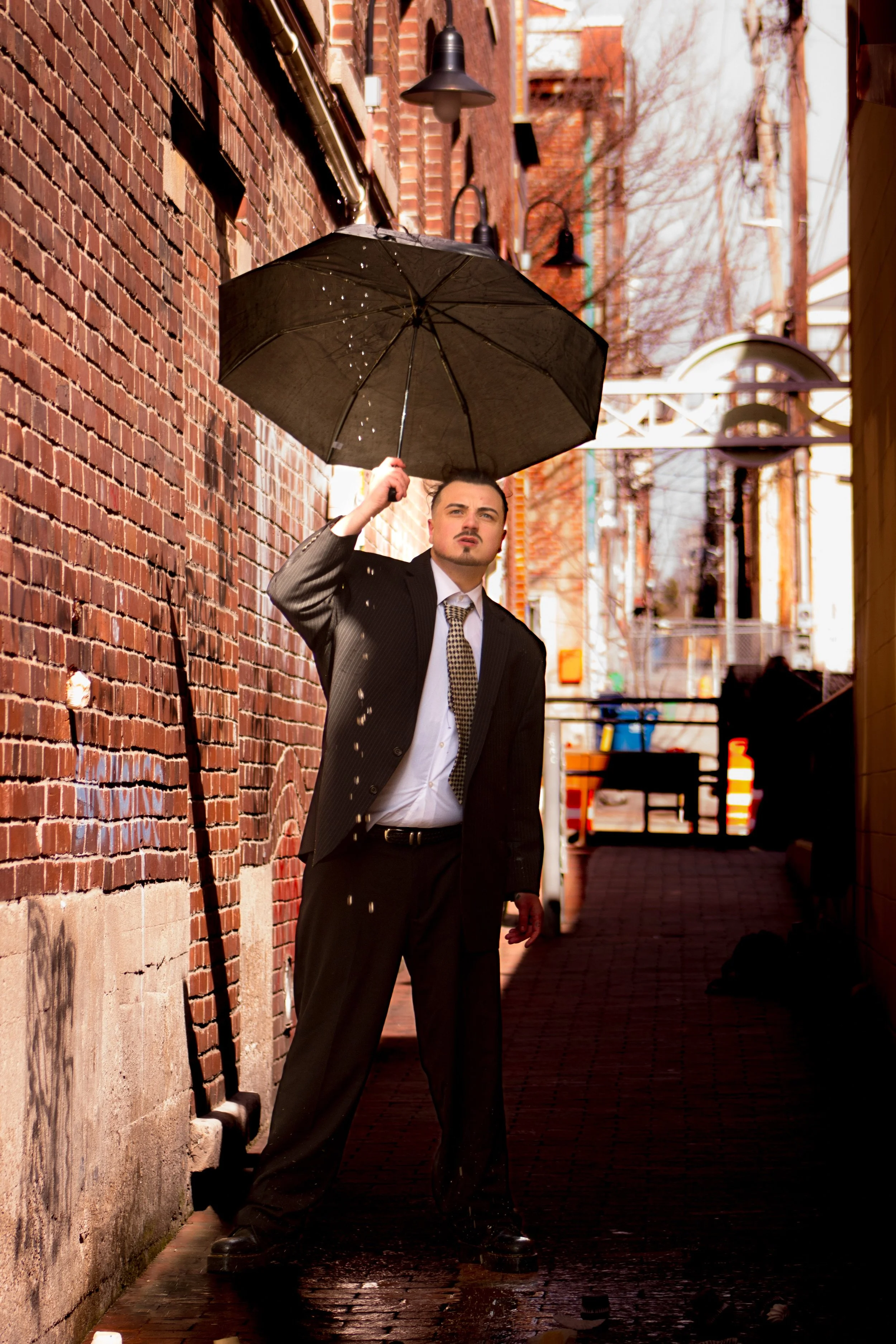 A man in a suit holding an open umbrella standing in a narrow alleyway on a rainy day.