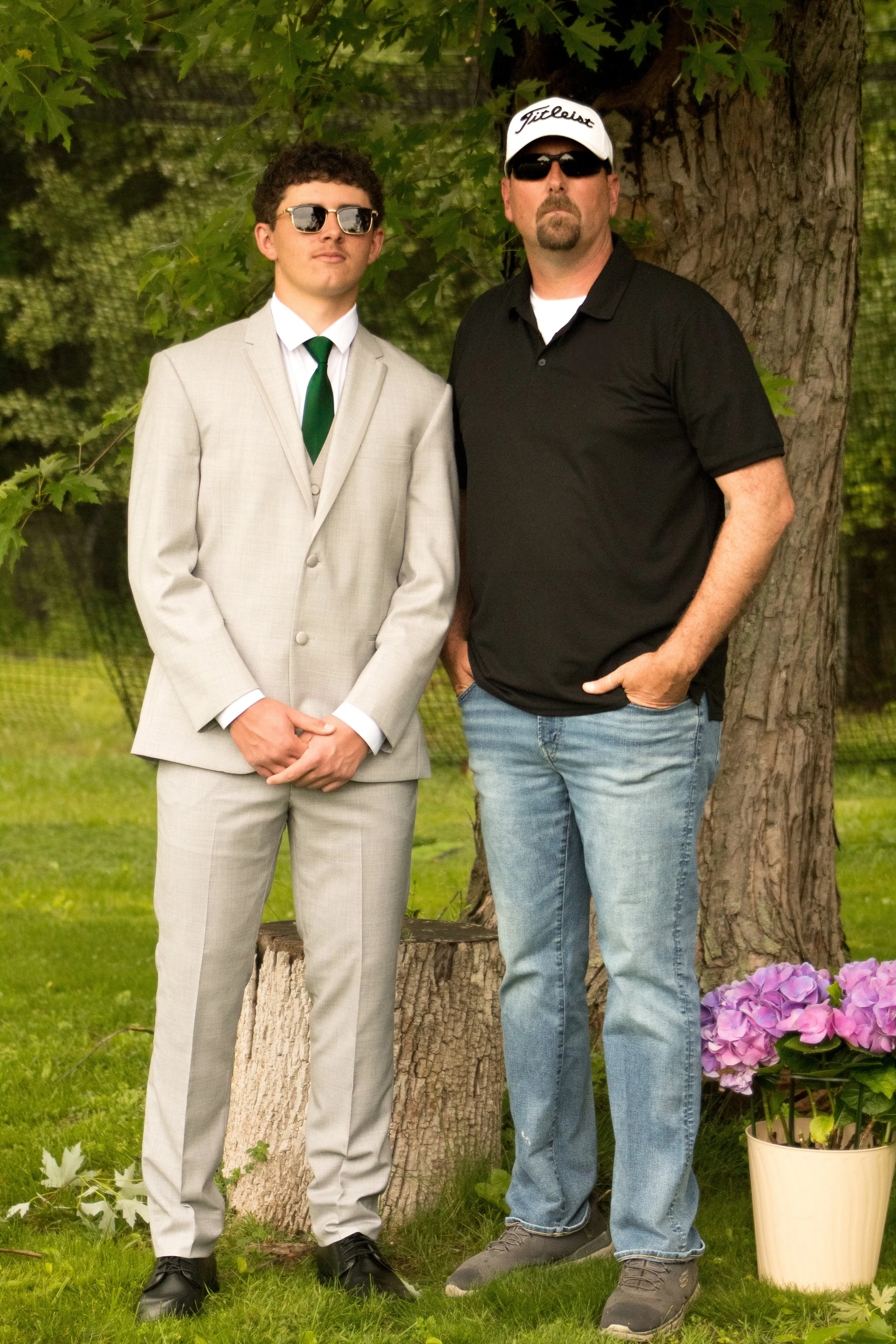 Two men standing outdoors near a large tree, with green foliage and purple hydrangea flowers in a beige pot nearby.