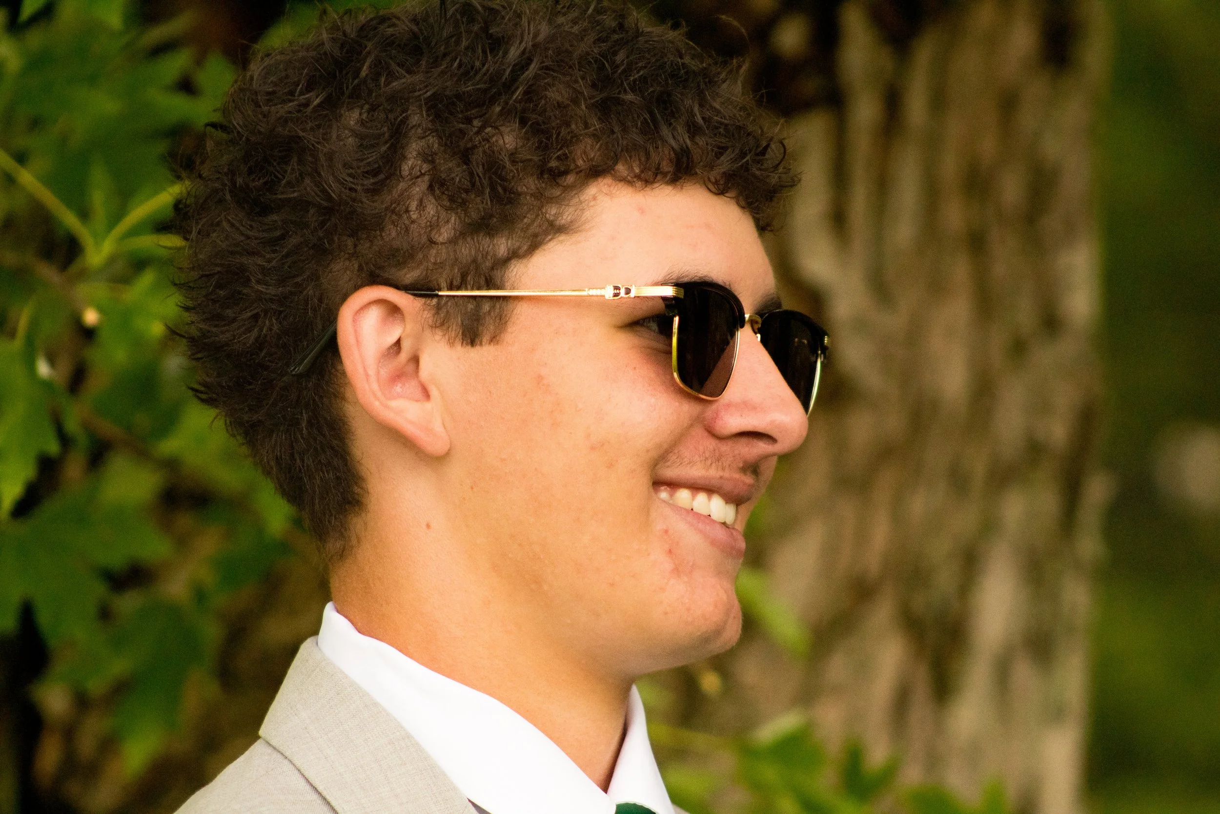 Close-up profile of a person with curly hair wearing sunglasses, a white shirt, and a light-colored suit jacket, smiling outdoors with green foliage in the background.