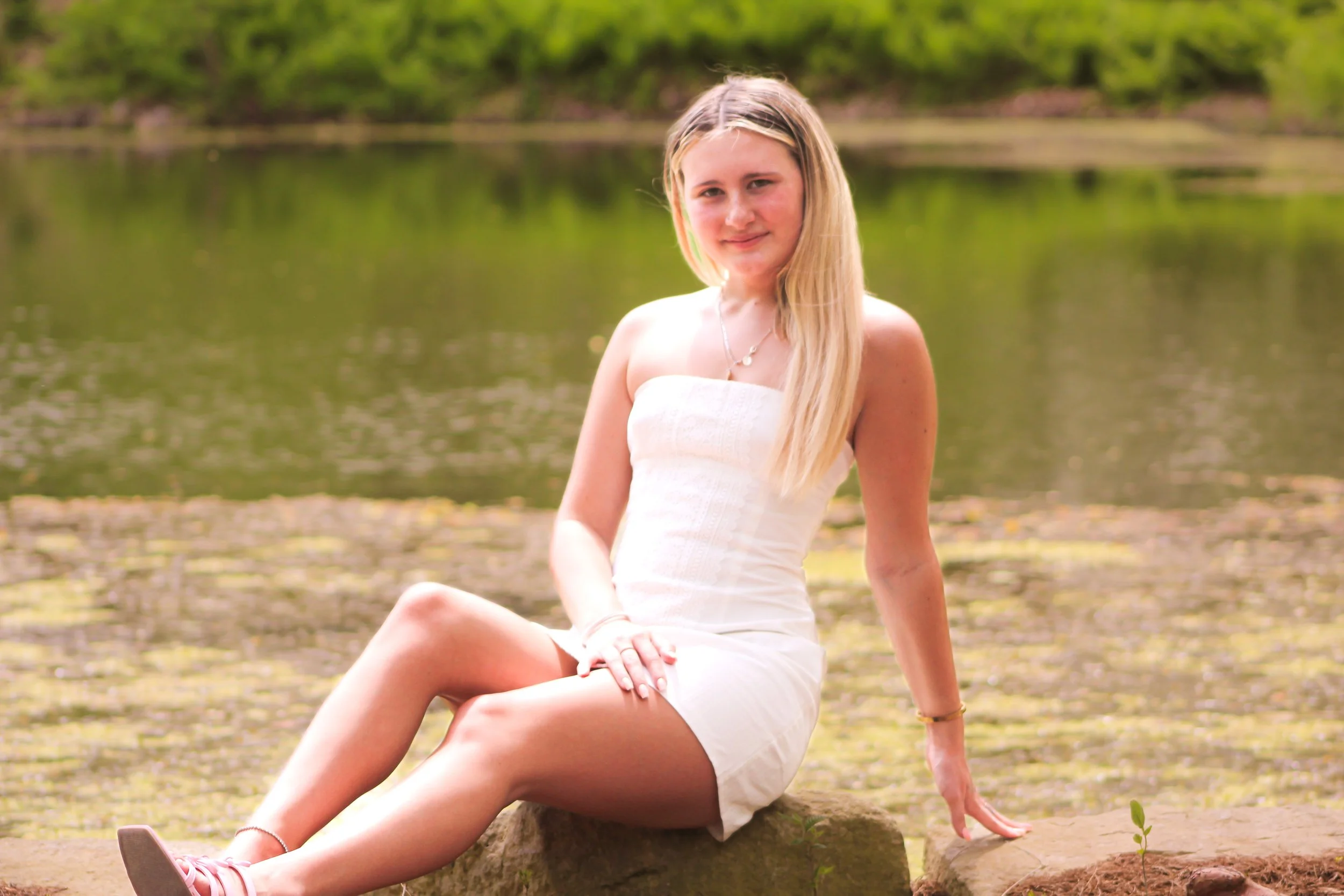 A young woman with long blonde hair sitting on a rock in front of a lake with green trees in the background, wearing a white strapless dress.