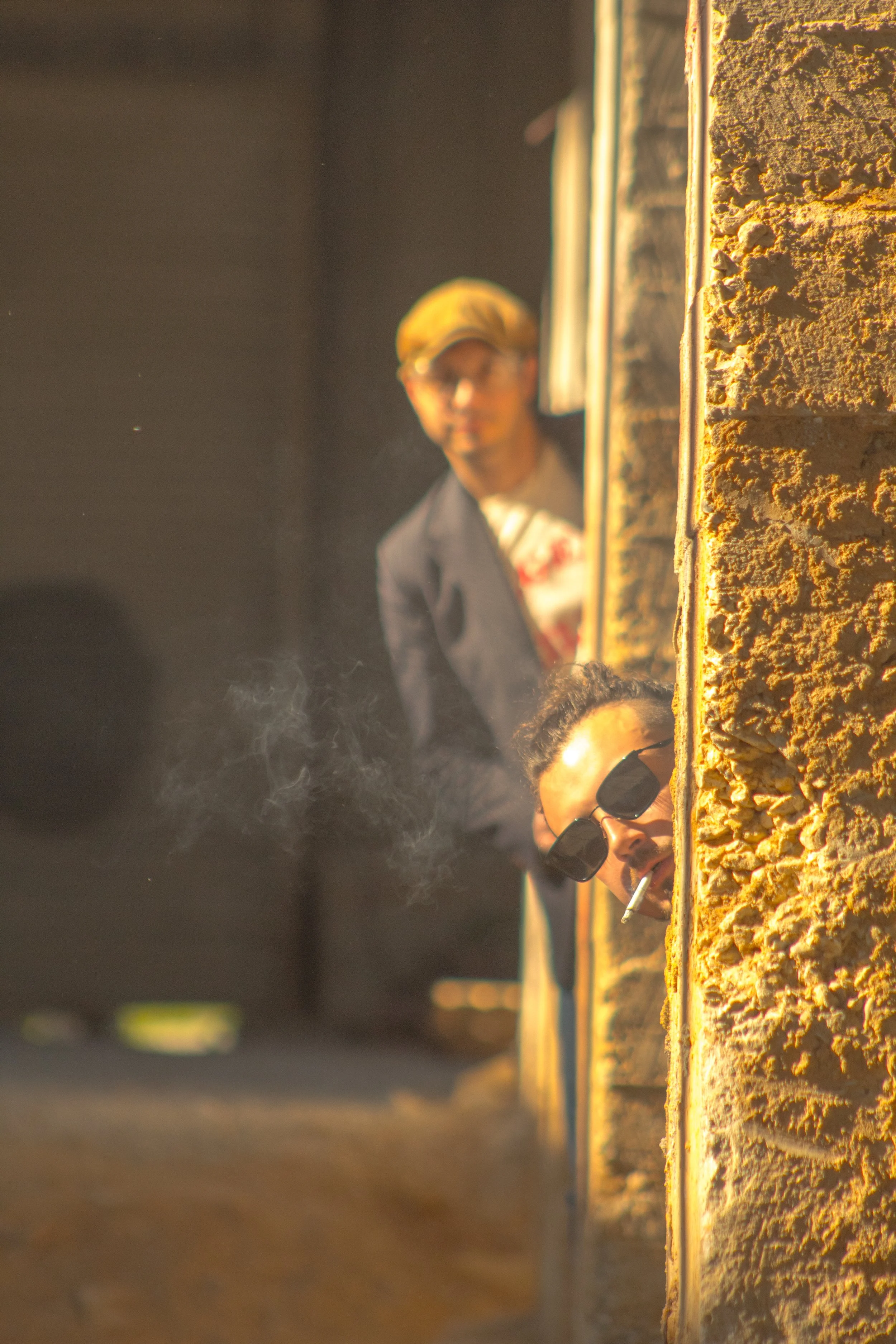 Three people peeking around a wall, one woman in sunglasses smoking a cigarette, two men in the background wearing glasses, with a warm, golden sunlight highlighting the textured wall.