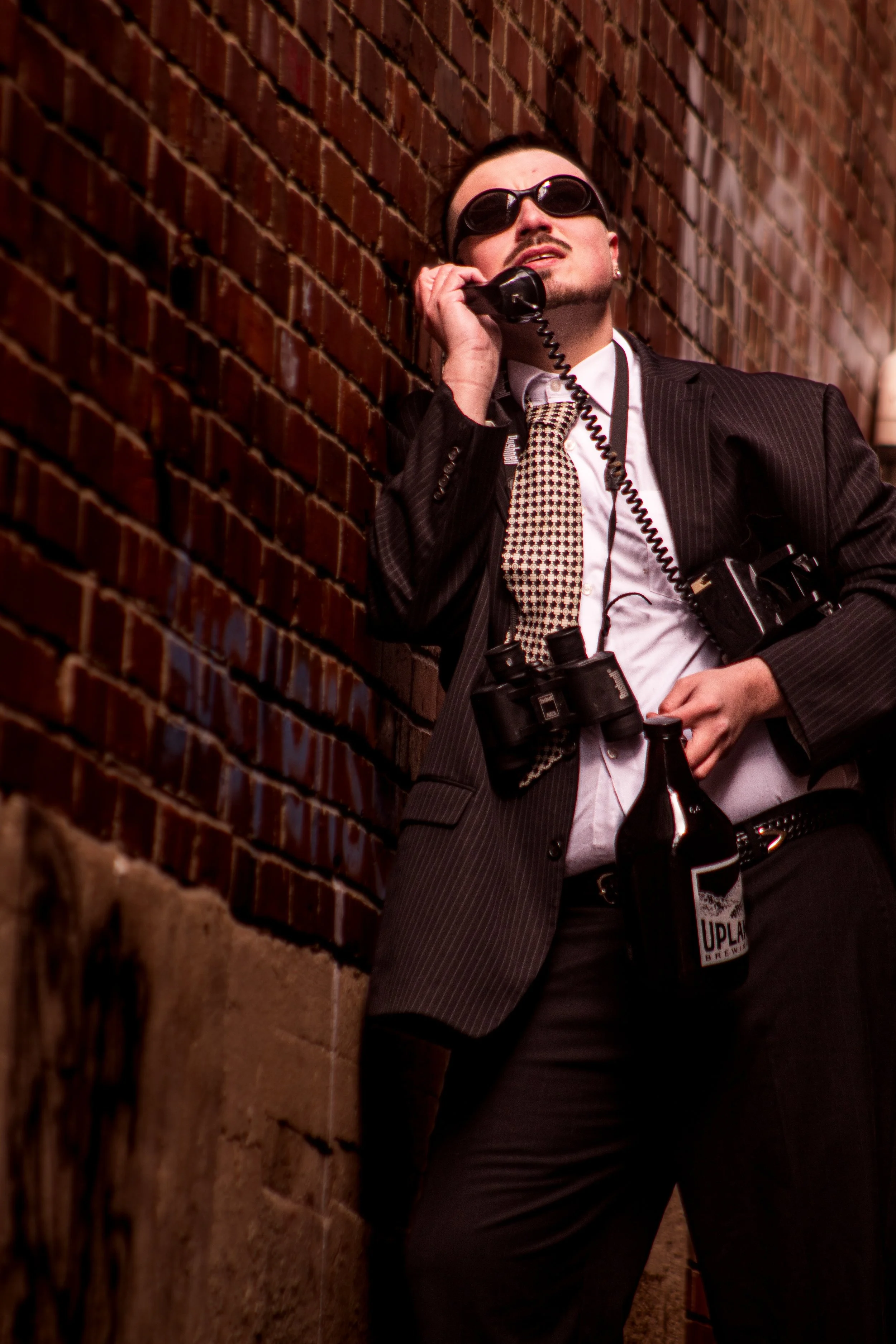 A man wearing sunglasses, a suit, and a patterned tie leaning against a brick wall while talking on a corded phone. He has binoculars around his neck, a camera hanging from his neck, and a beer bottle tucked into his belt.