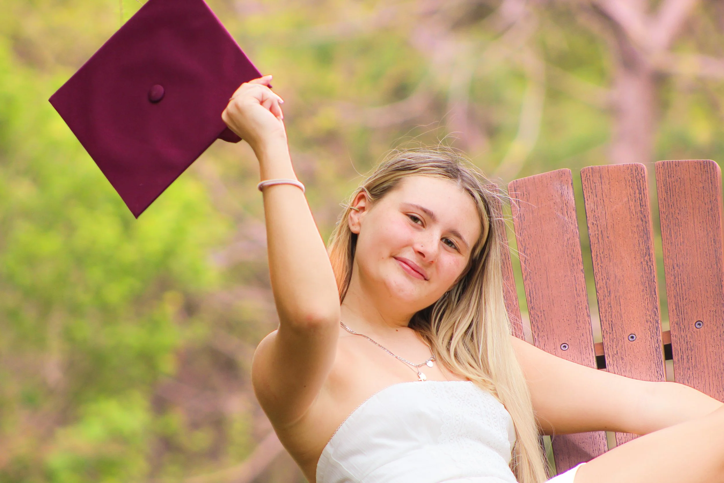 A young woman sitting outdoors on a wooden chair, holding a maroon graduation cap, smiling at the camera with a background of trees and greenery.