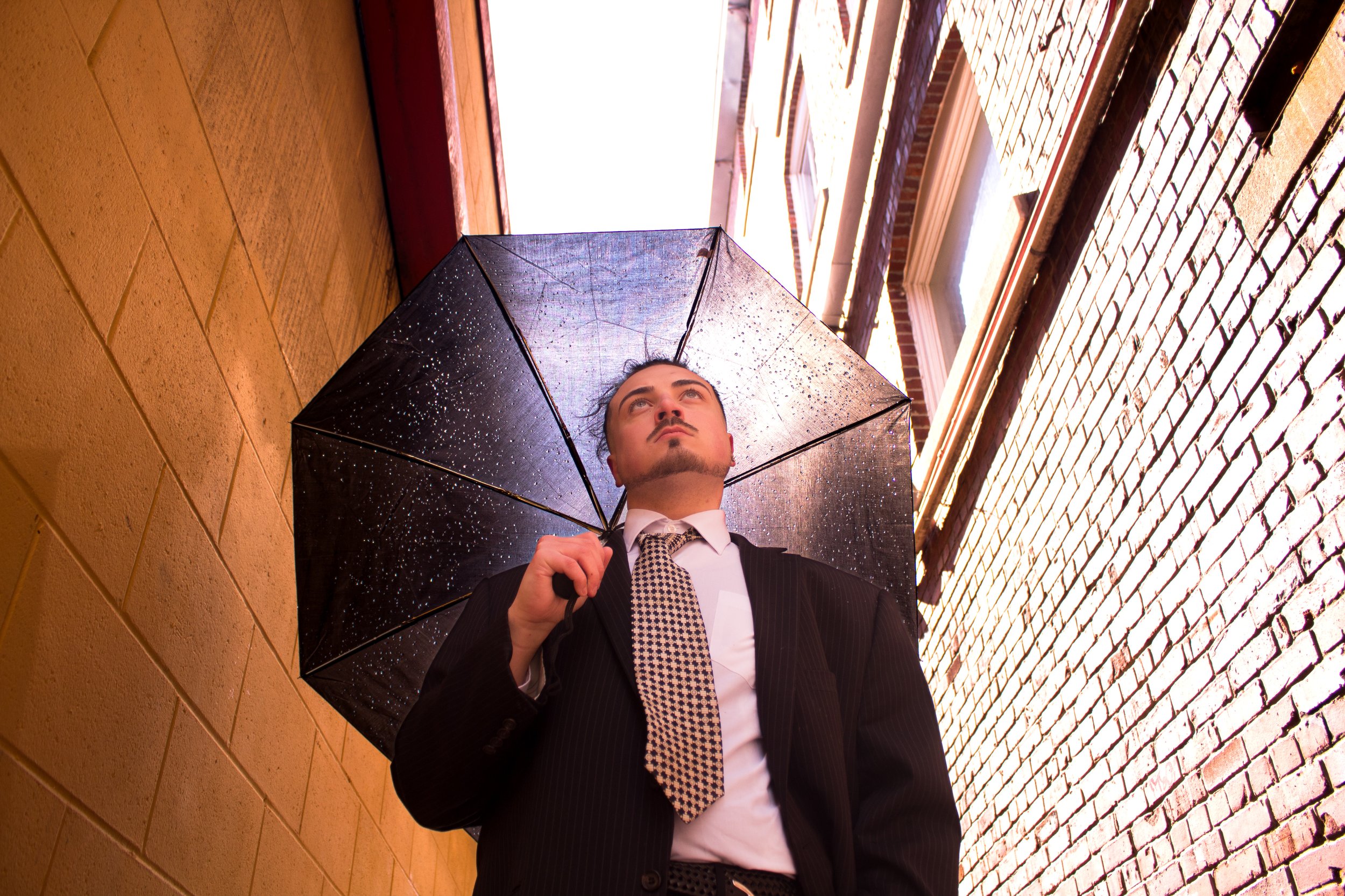 A man in a black suit and tie holding a black umbrella with rain droplets, standing in a narrow alley between brick buildings.