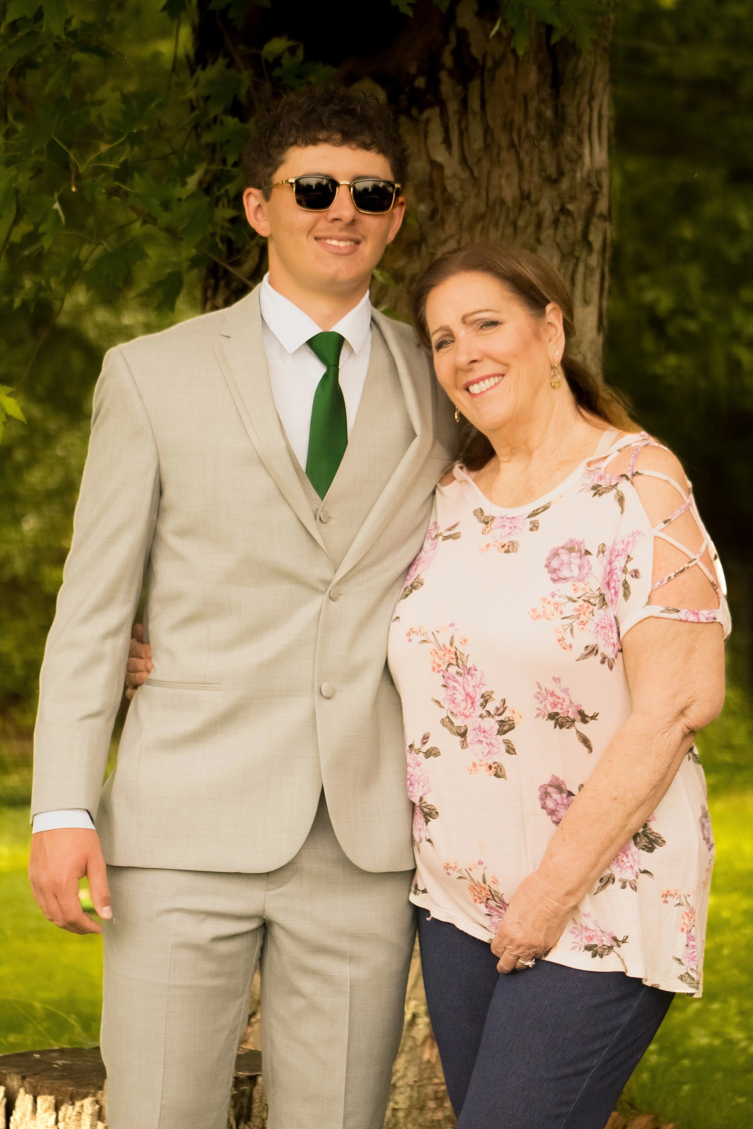 A young man wearing sunglasses, a beige suit, and a green tie standing next to a woman with brown hair, wearing a floral top and dark pants, both smiling outdoors near a tree.
