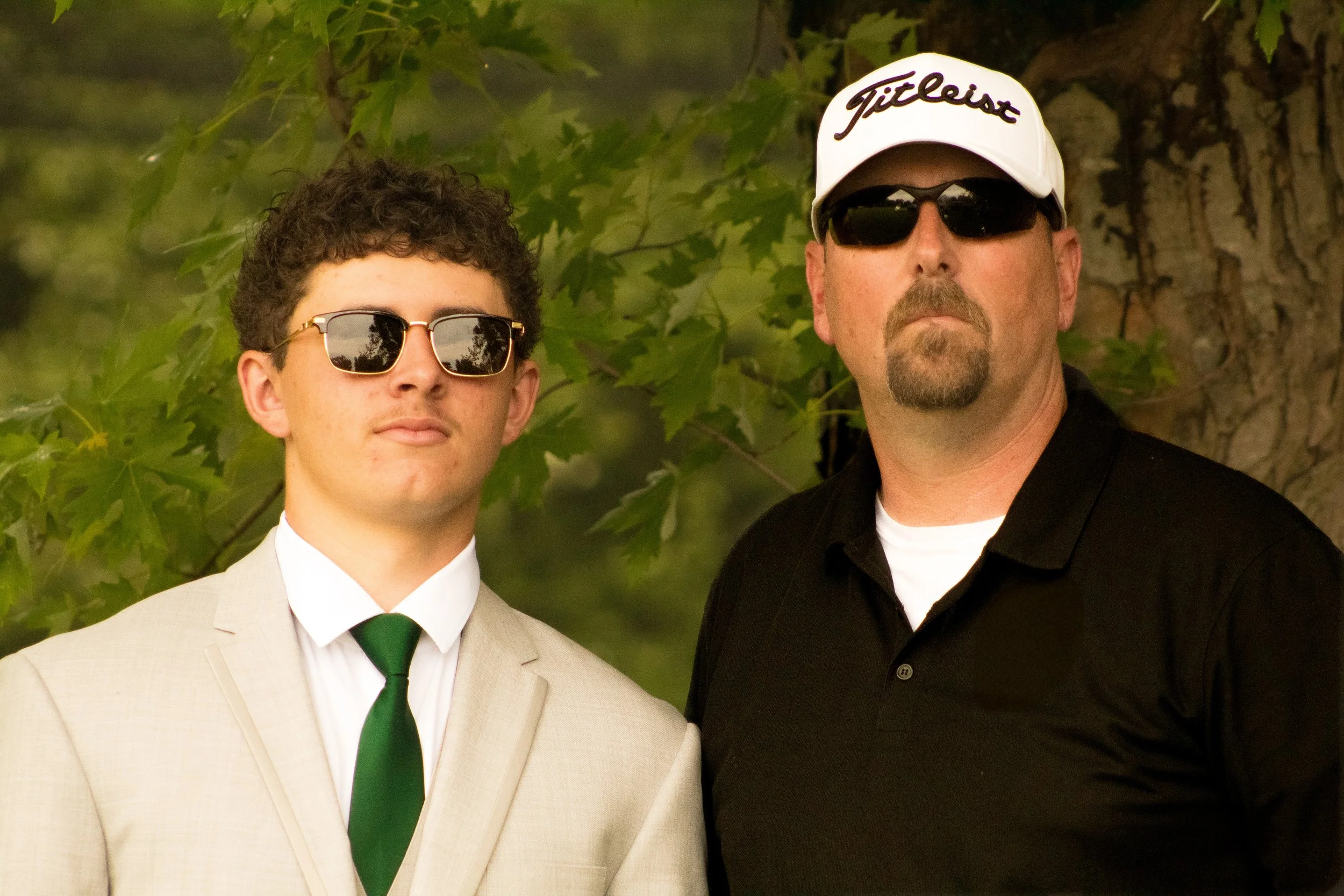 Two men standing outdoors, one young man in a beige suit with a green tie and dark sunglasses, and an older man in a black shirt with a white Titleist cap and dark sunglasses, both looking at the camera.
