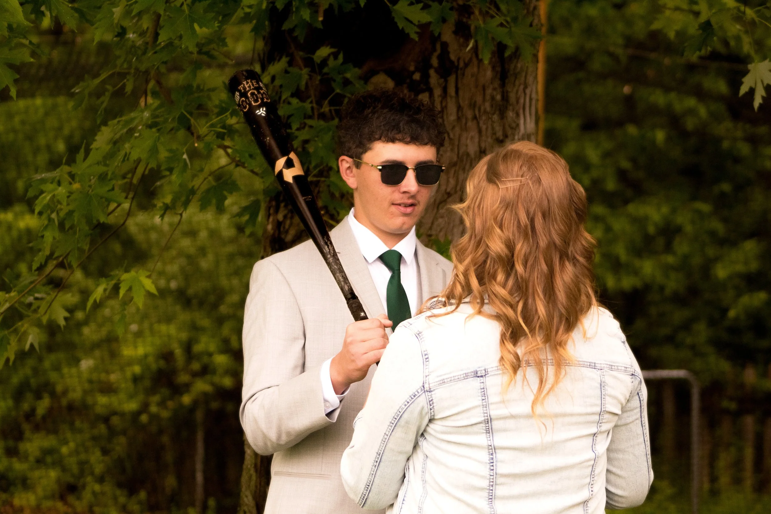 A young man in sunglasses and a light-colored suit stands outdoors near a tree, holding a baseball bat, while a woman with long, wavy red hair faces him, in a wooded area.
