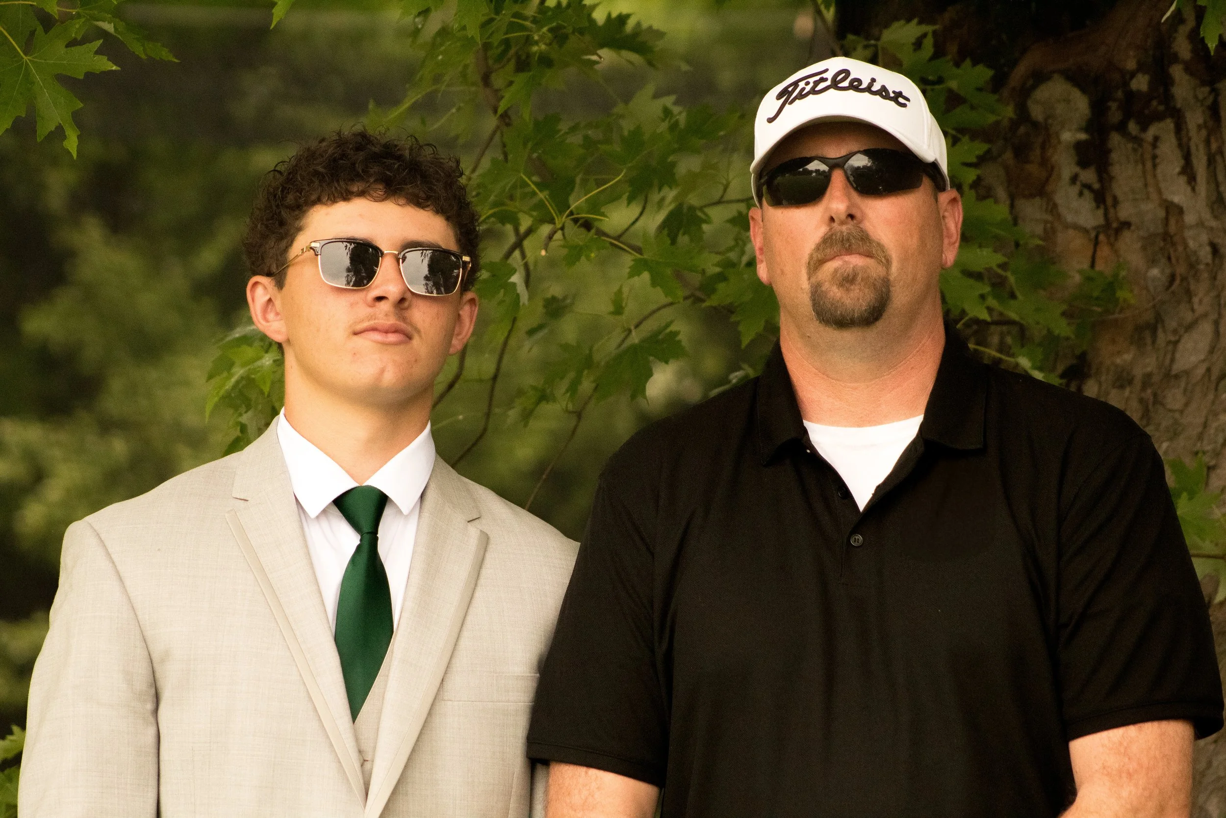 Two men standing outdoors surrounded by green leaves. The man on the left is wearing a light-colored suit, white shirt, green tie, and sunglasses. The man on the right is wearing a black shirt, a white cap with 'Titleist' written on it, and sunglasse