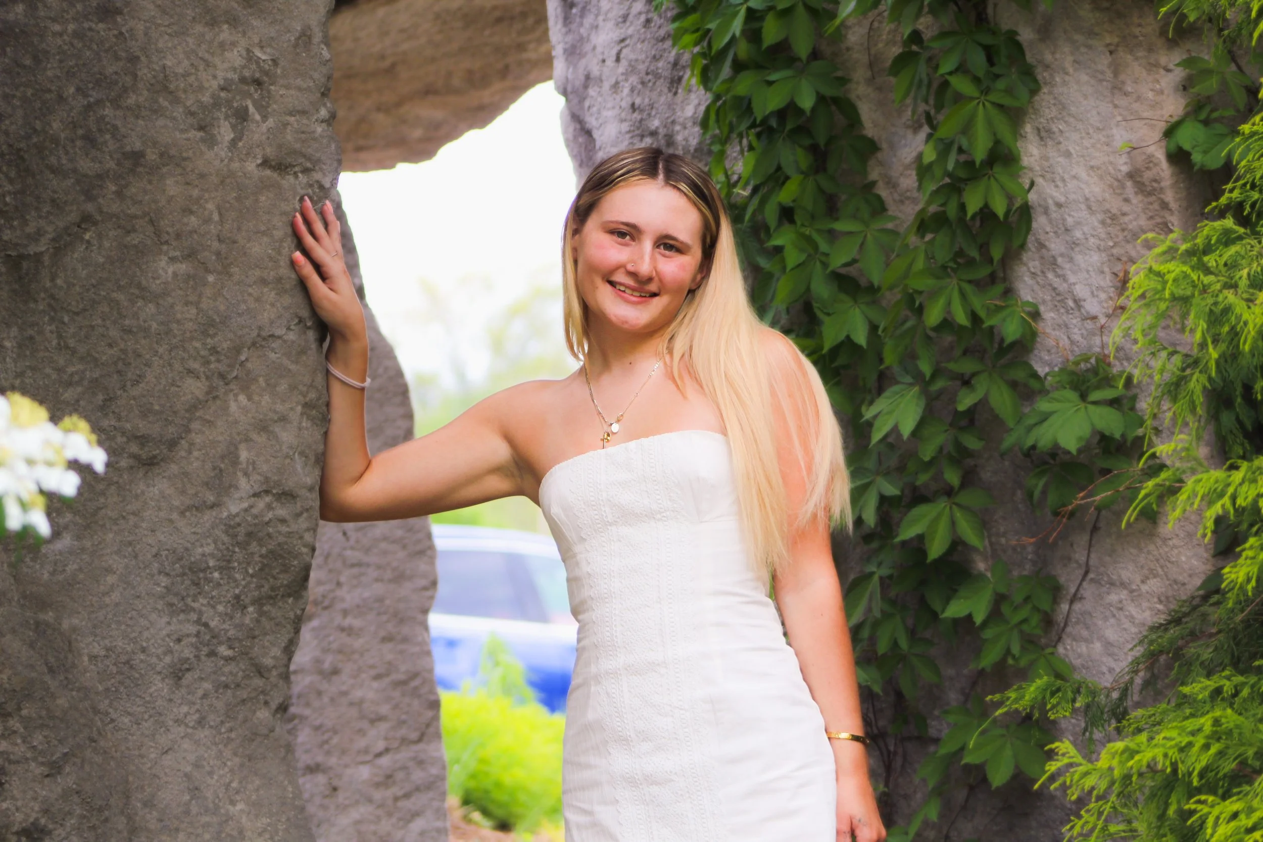 A young woman with long blonde hair, wearing a white strapless dress, standing between two large rocks with green foliage behind her, smiling at the camera.