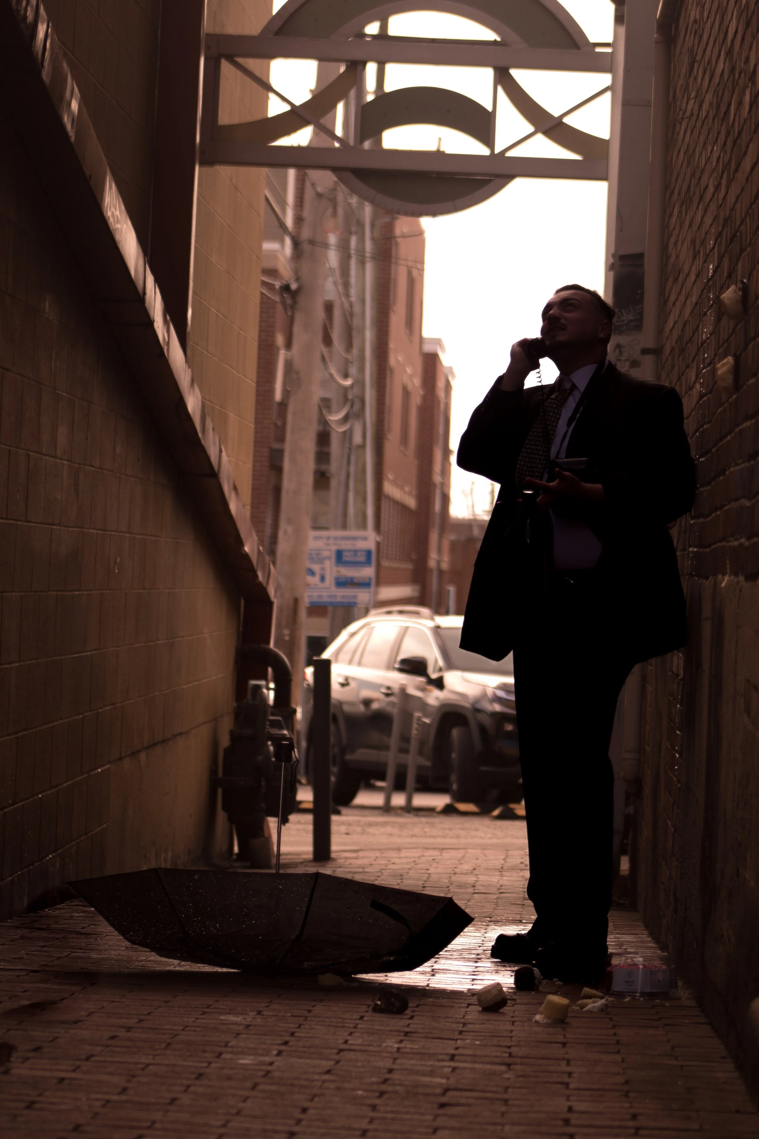 A silhouette of a man wearing a suit, talking on a mobile phone while standing in a narrow alley with an umbrella on the ground and some scattered trash.