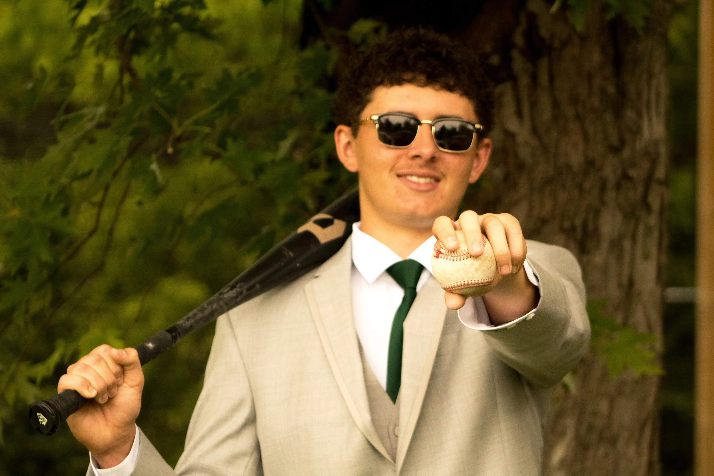 A man in a beige suit and sunglasses holding a baseball while standing outdoors near a tree.