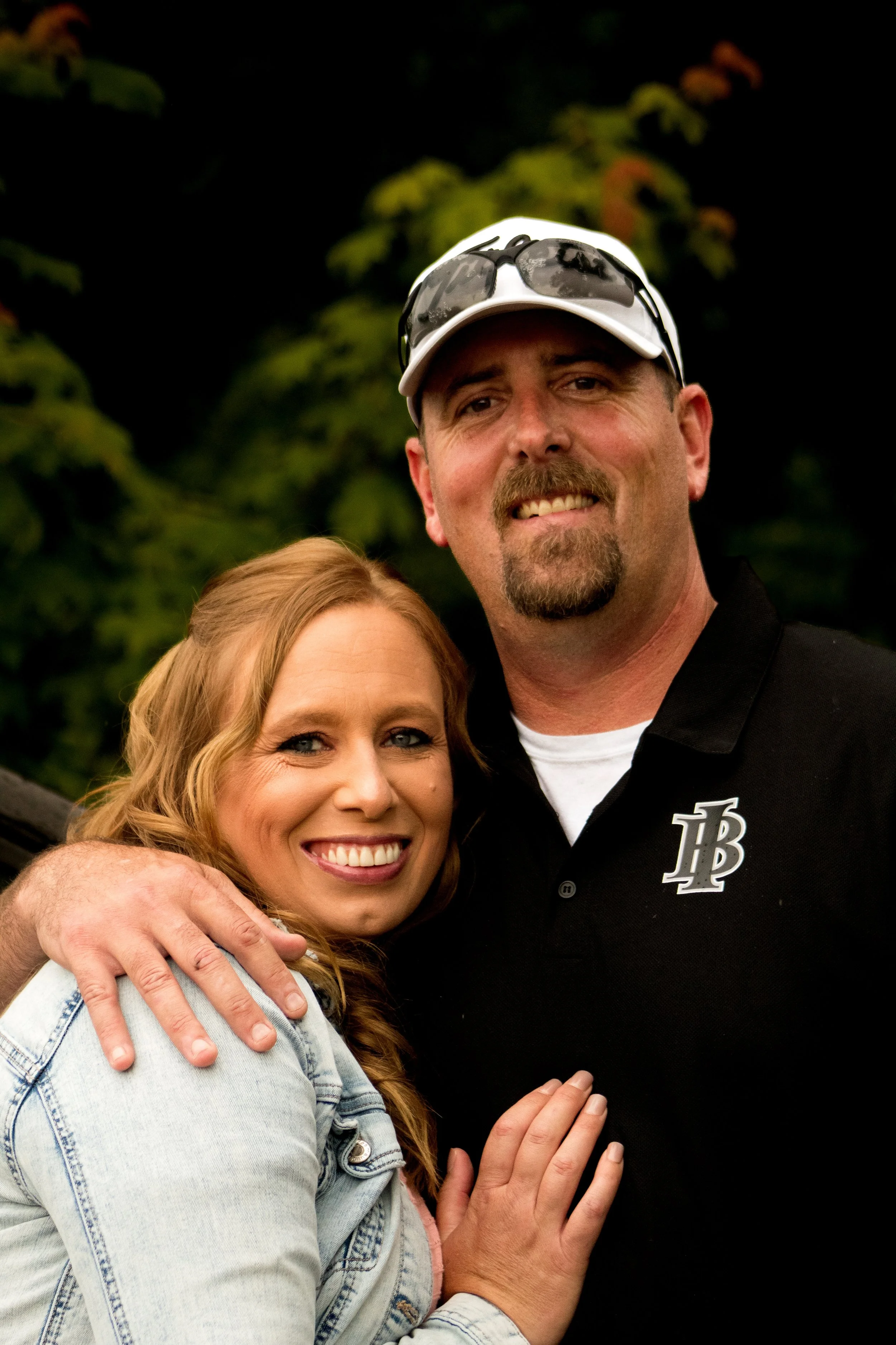 A man with a goatee and sunglasses on his white cap, smiling, standing next to a woman with red hair, smiling, both embracing with a background of trees.
