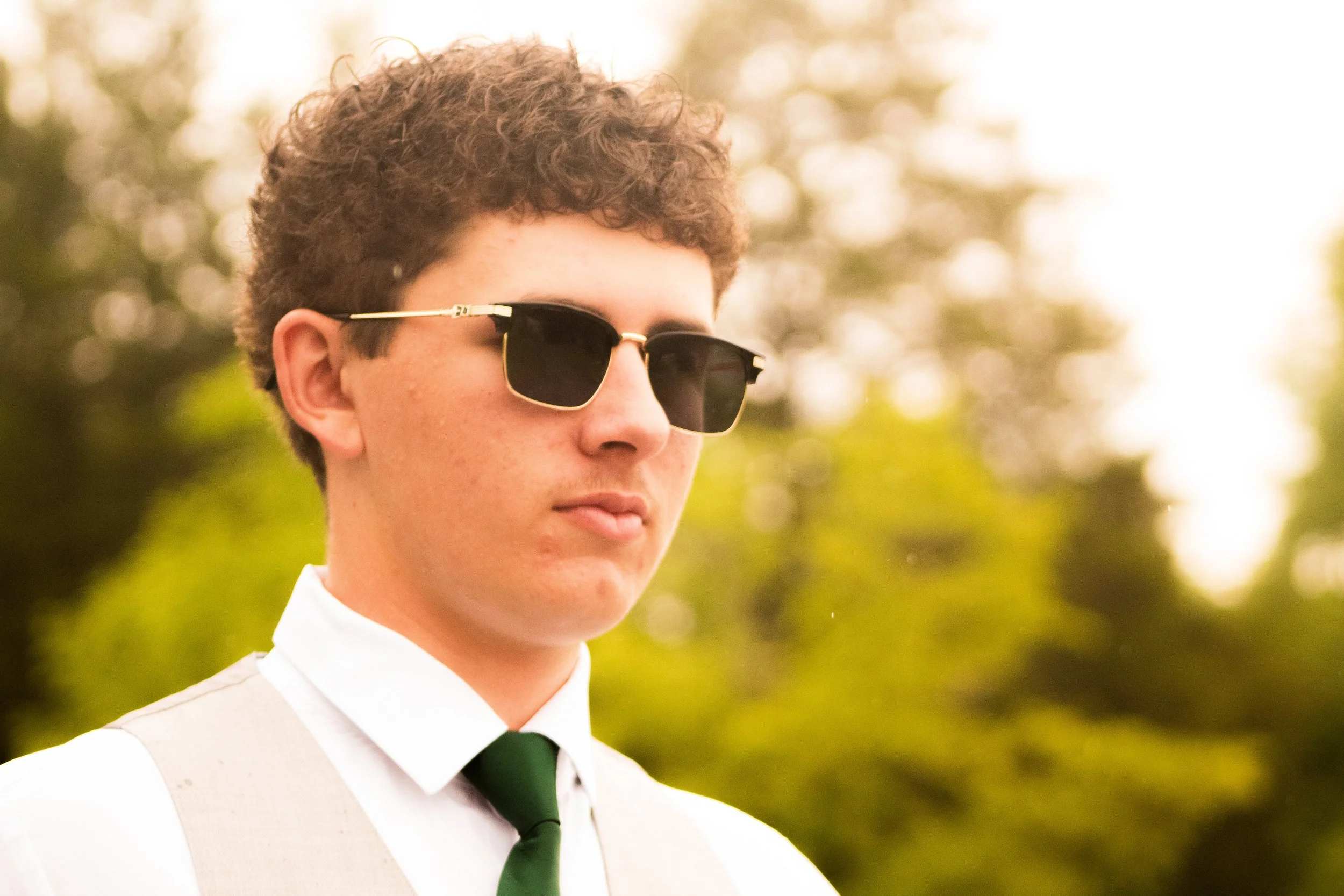 Young man in formal attire, wearing sunglasses, white dress shirt, beige vest, and green tie, outdoors with blurred trees in the background.