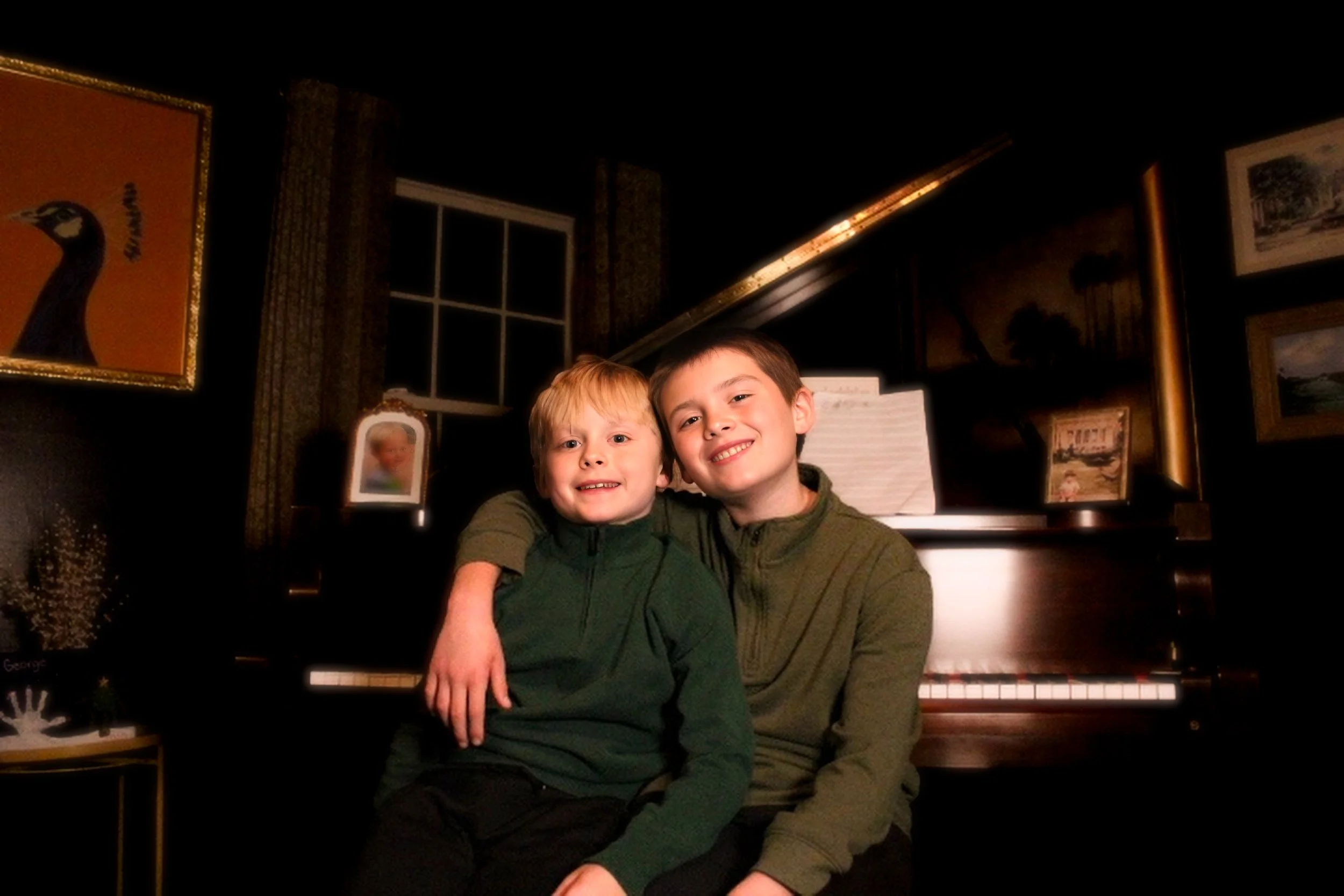 Two boys sitting close together in front of a piano, smiling, with warm indoor lighting, dark walls, and framed artwork on the wall.