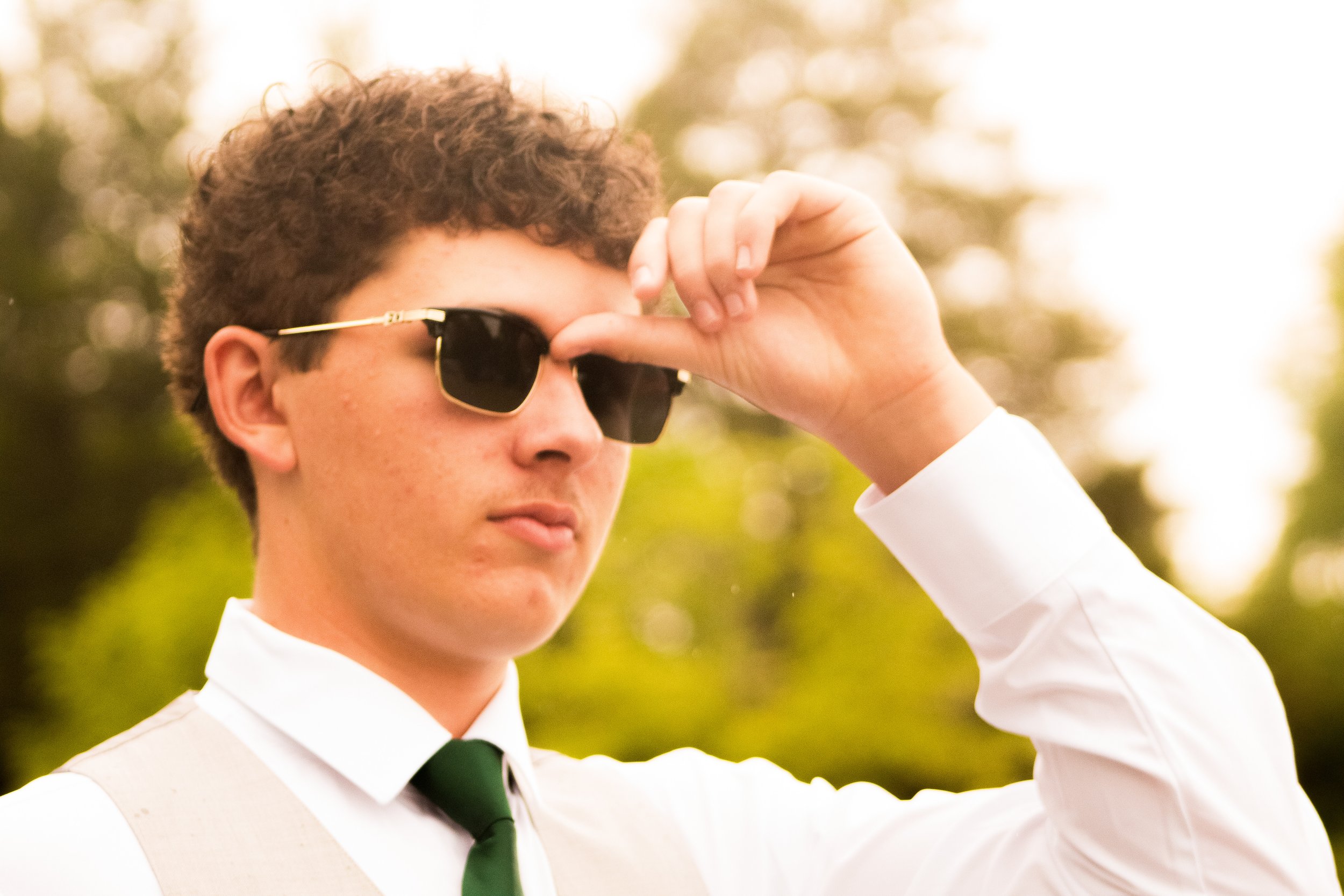 A young man wearing sunglasses and a white shirt with a green tie, adjusting his sunglasses outdoors during daytime with a blurred background of trees and sunlight.