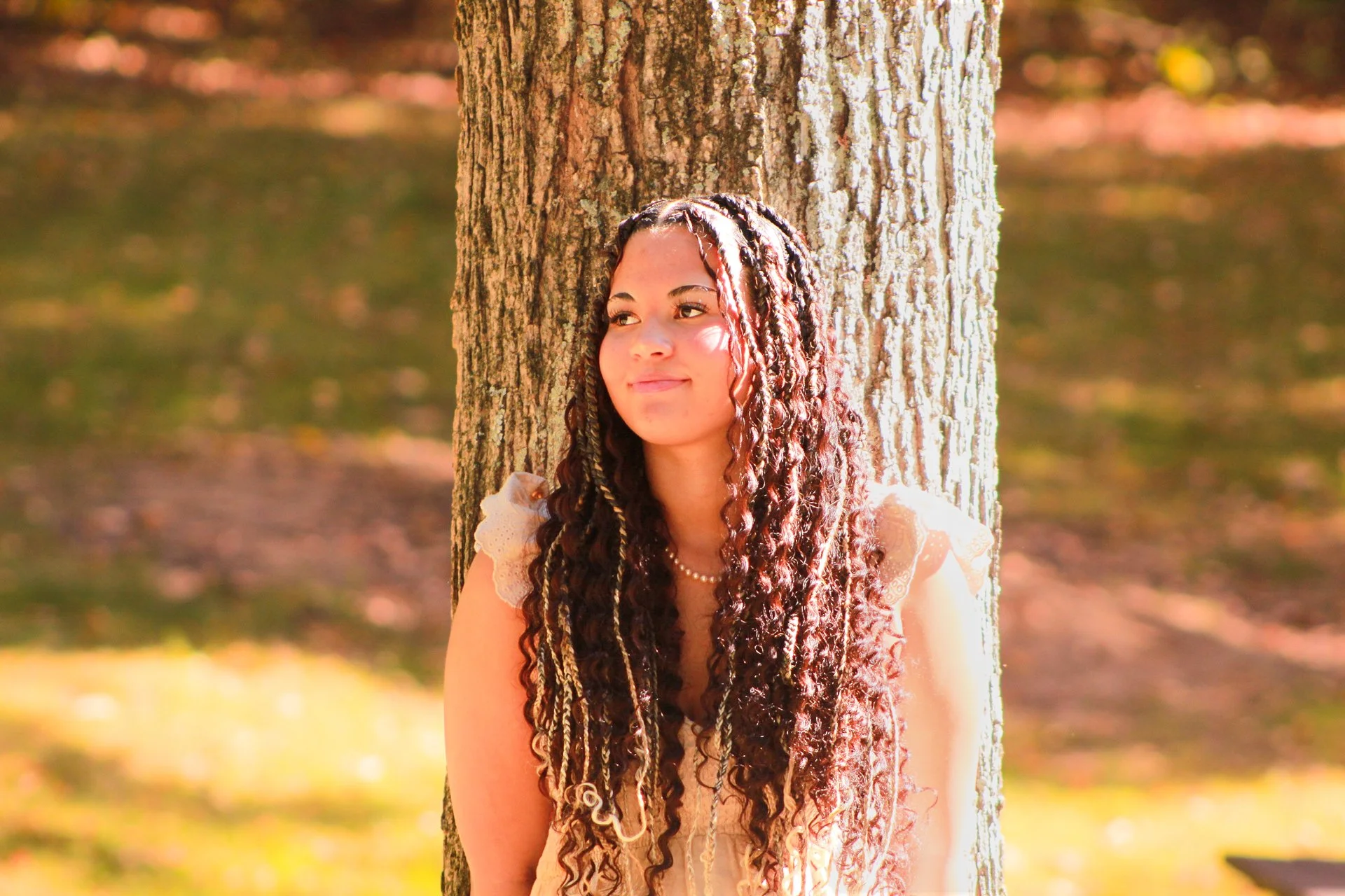 A young woman with long, curly hair standing against a tree outdoors on a sunny day, looking thoughtfully to the side.
