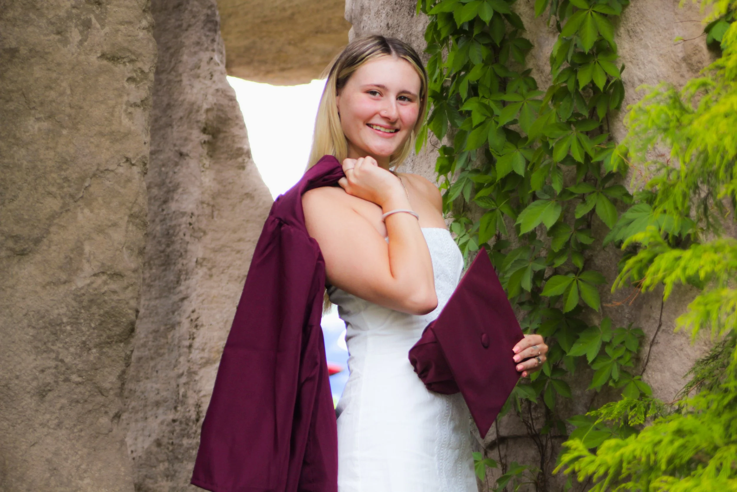 Young woman in a white dress holding a maroon graduation cap and gown, standing between stone archways with green ivy, smiling for a photo.