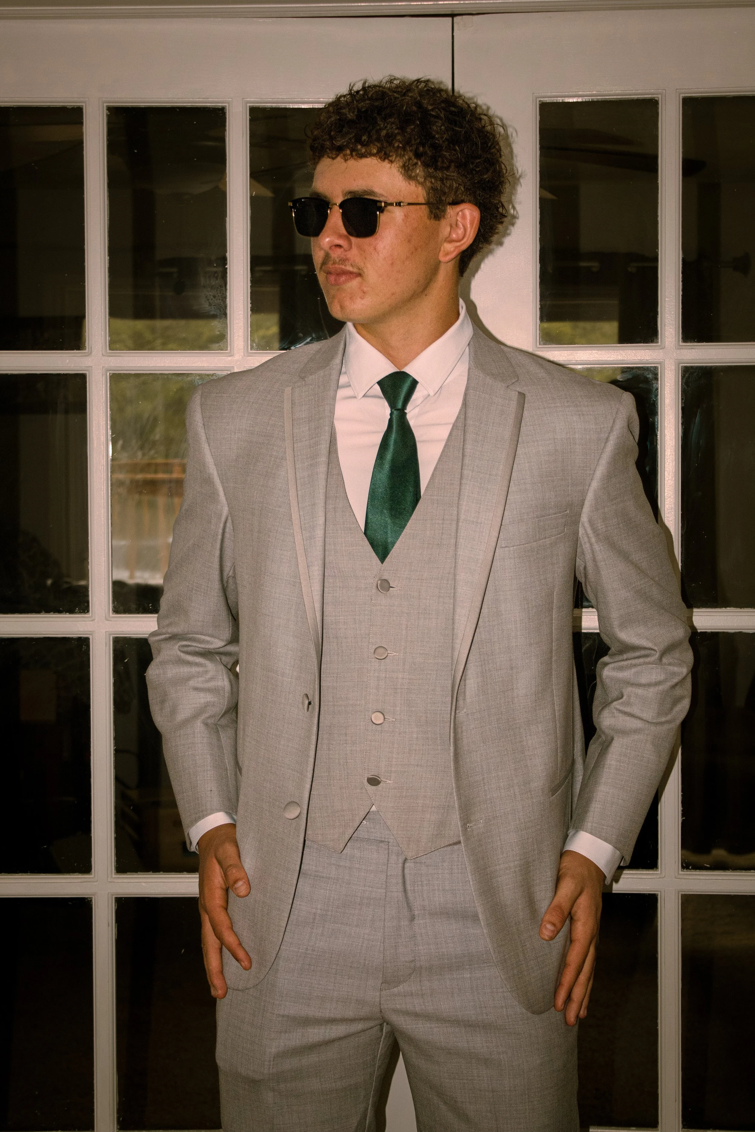 Man in a light gray suit, green tie, white shirt, wearing black sunglasses, standing in front of glass-paneled door.