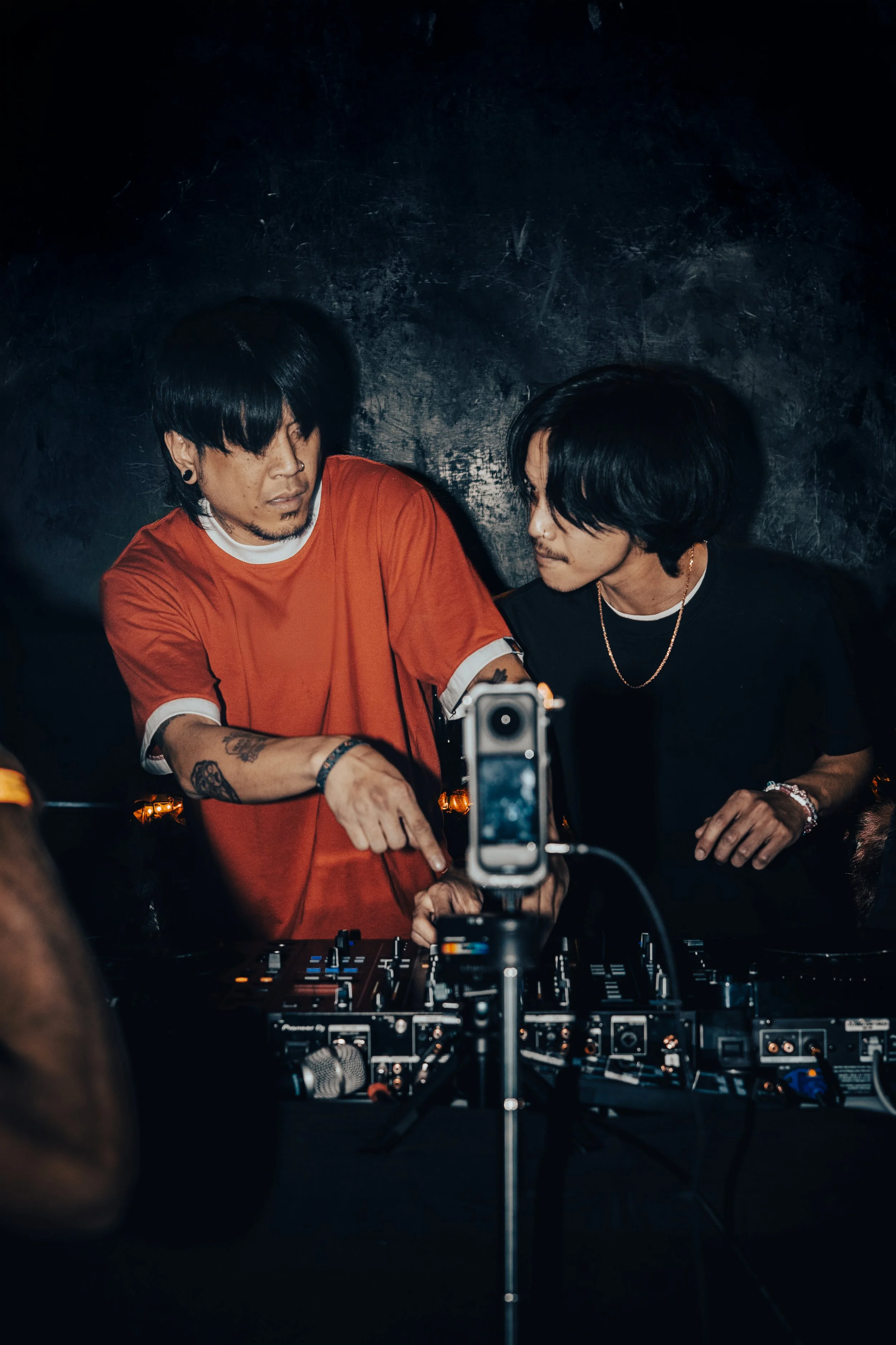 Two DJs sit behind a deck in a darkly lit event setting. A 360 video camera is seated in front of them.