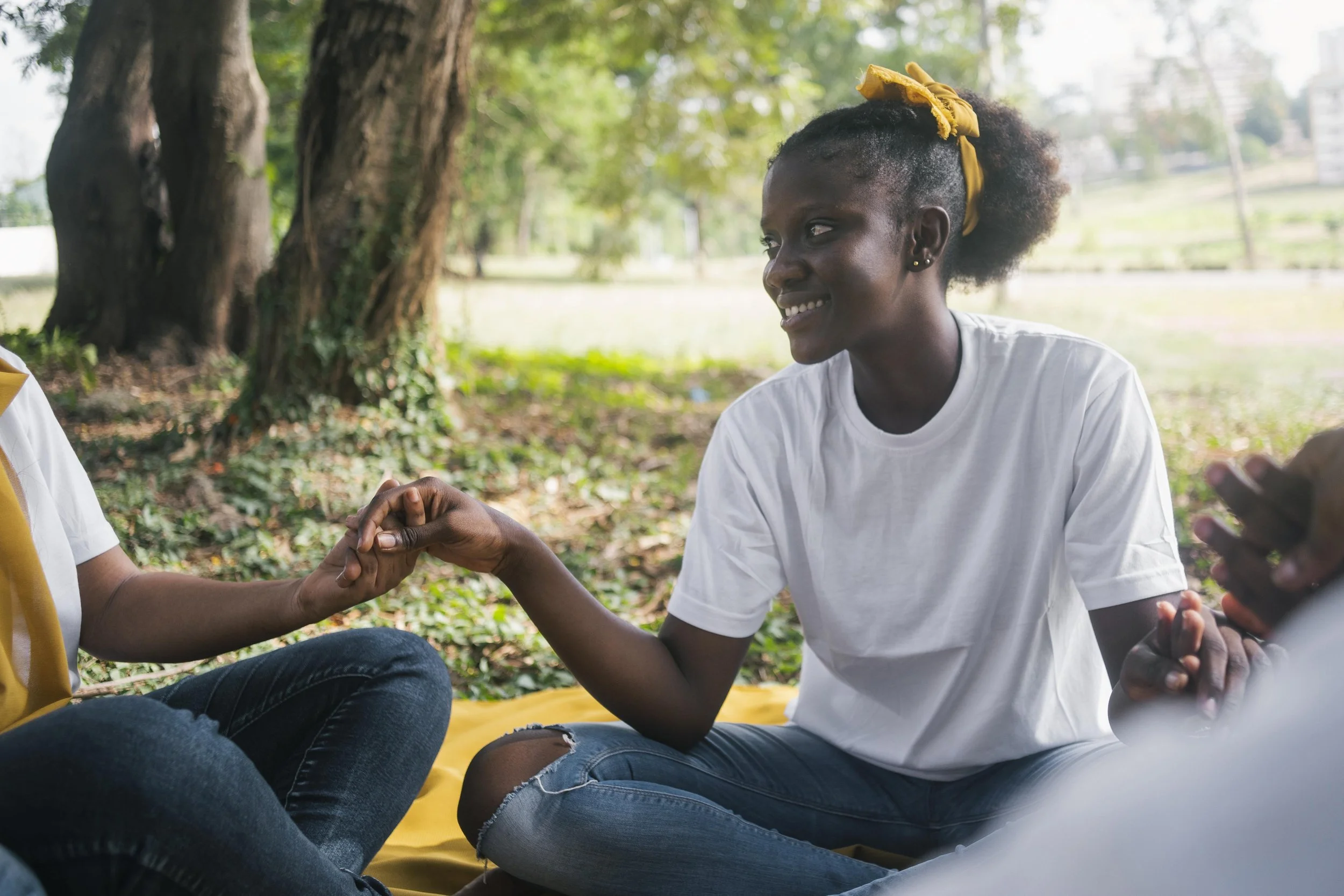 A young woman with dark curly hair tied with a yellow scarf smiling and holding hands with someone outdoors, sitting on a yellow blanket in a park with green trees.