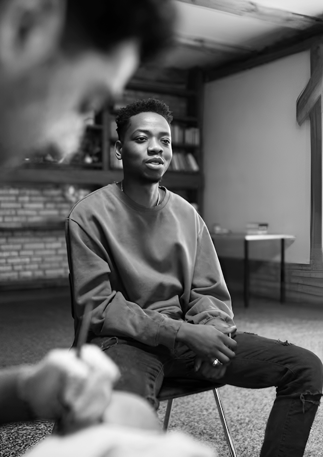 A young man sitting on a chair and looking attentively during a conversation in a cozy room with wood paneling and a bookshelf.